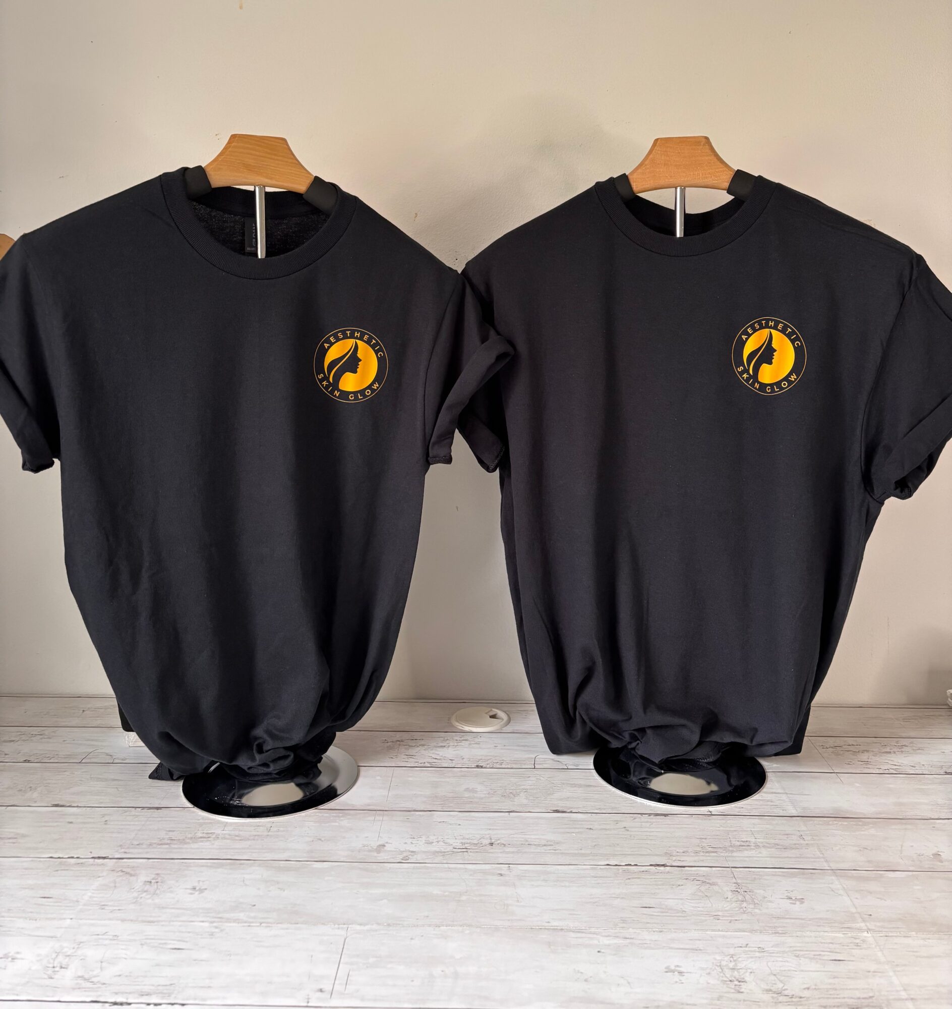 Two black t-shirts with a yellow and black circular logo on the chest, hanging on wooden hangers against a light wall.
