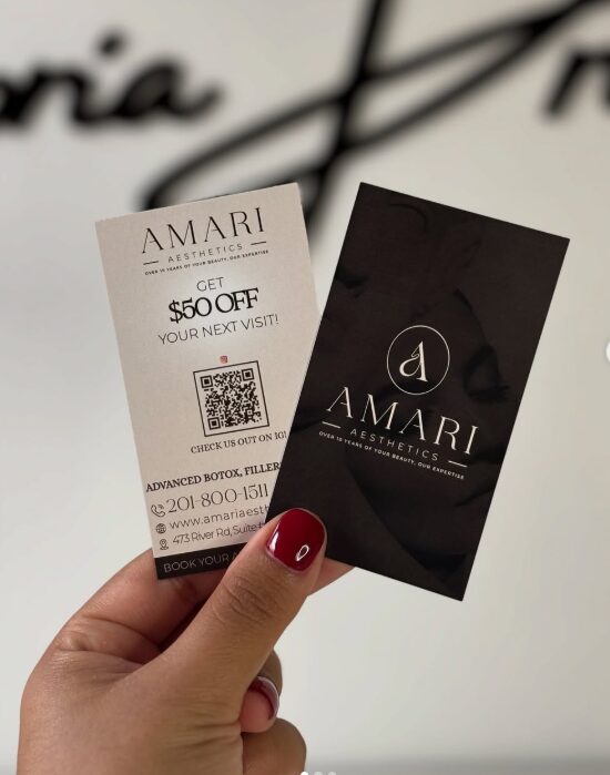 Person holding two business cards for Amari Aesthetics, one white with a QR code, one black with logo, against a blurred background.