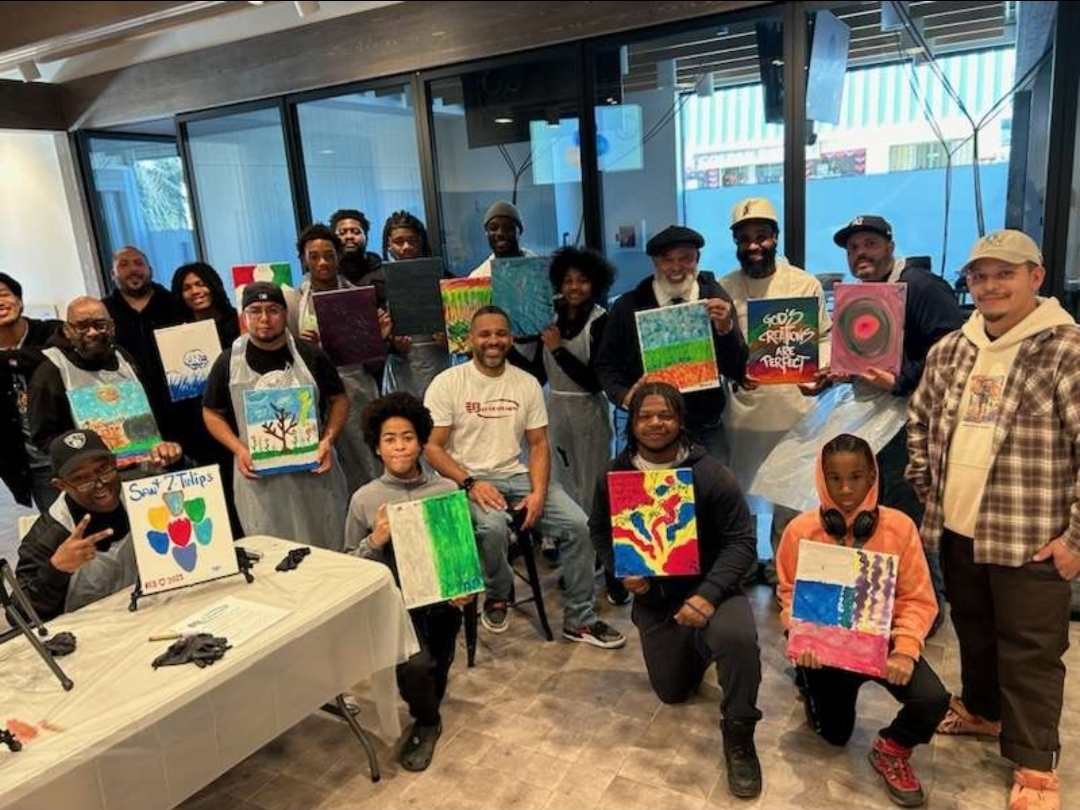 Group of people holding colorful paintings indoors with large windows in the background.