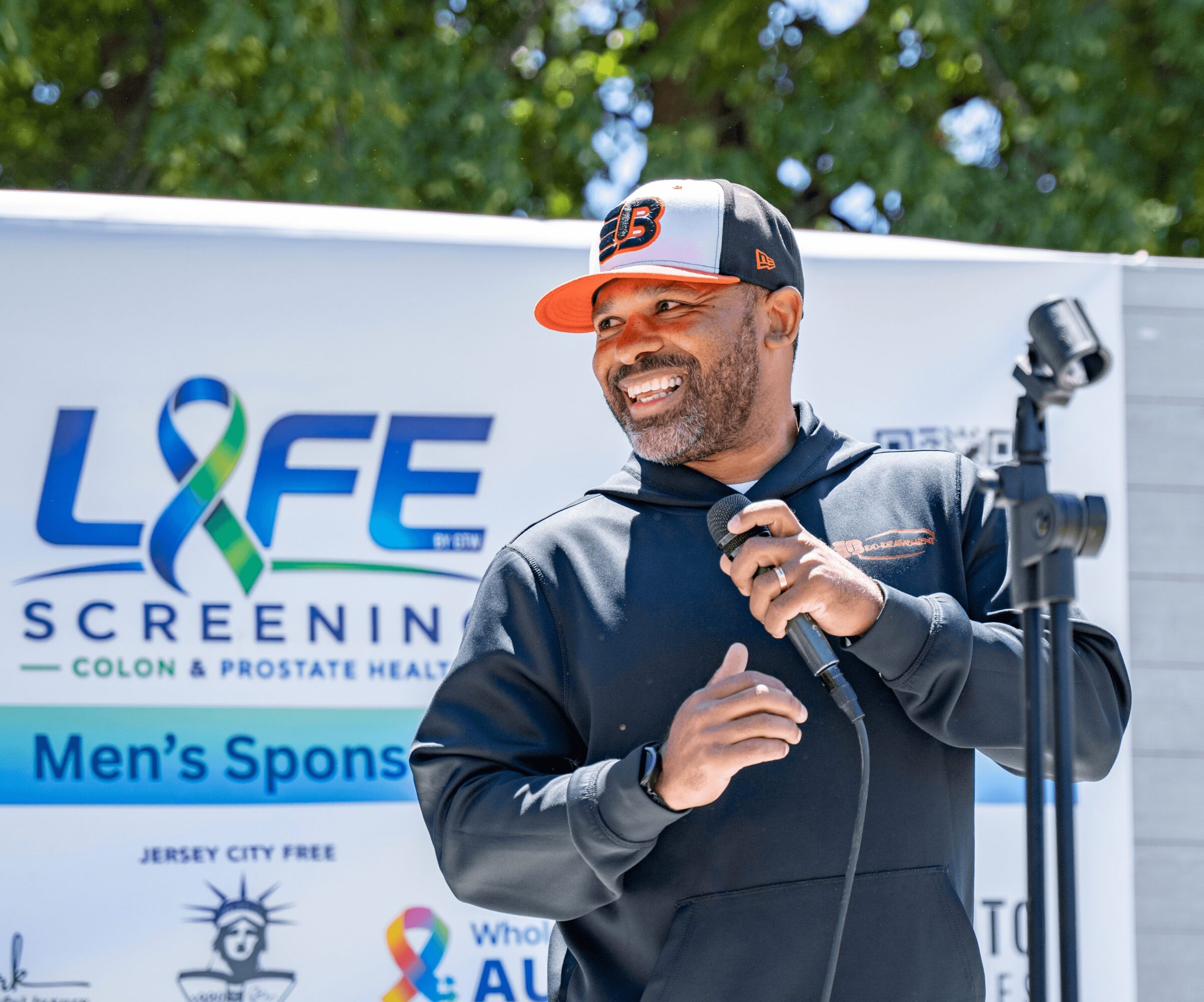 Man smiling, wearing a cap and jacket, standing outdoors near a banner with the word 'LIFE' and a ribbon symbol.