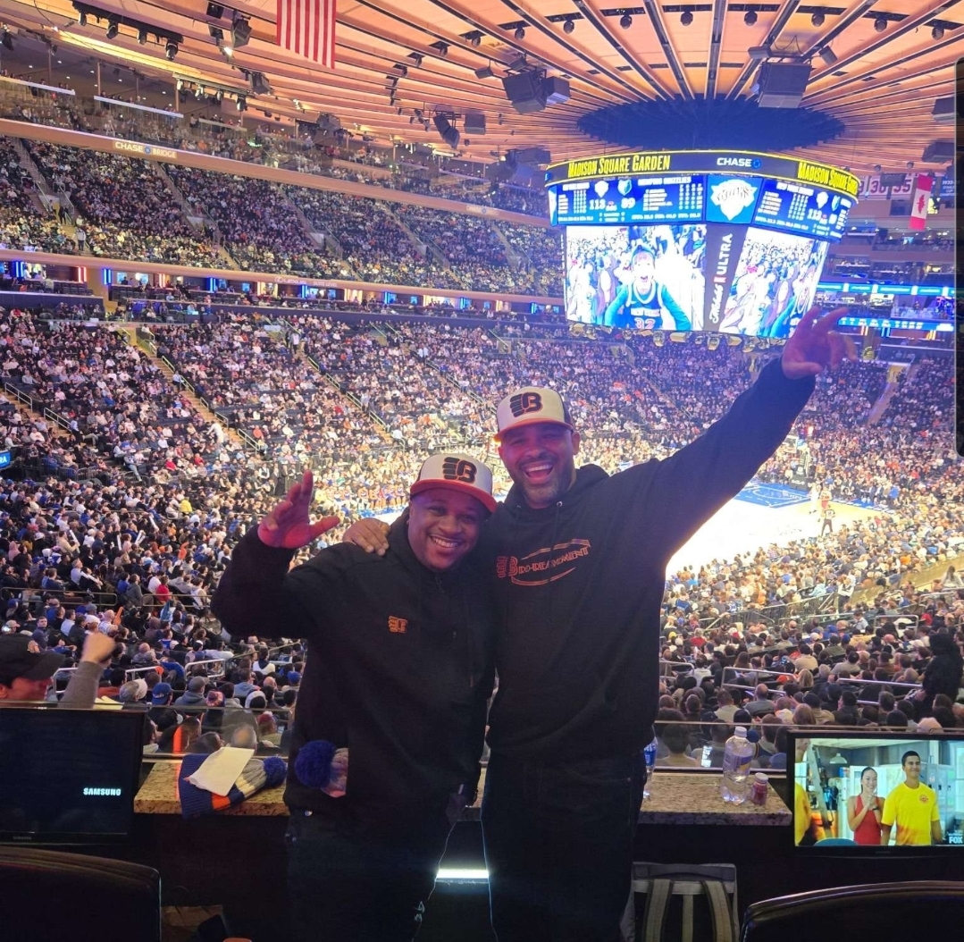 Two people smiling and posing with arms raised inside a large sports arena filled with spectators and a bright scoreboard overhead.