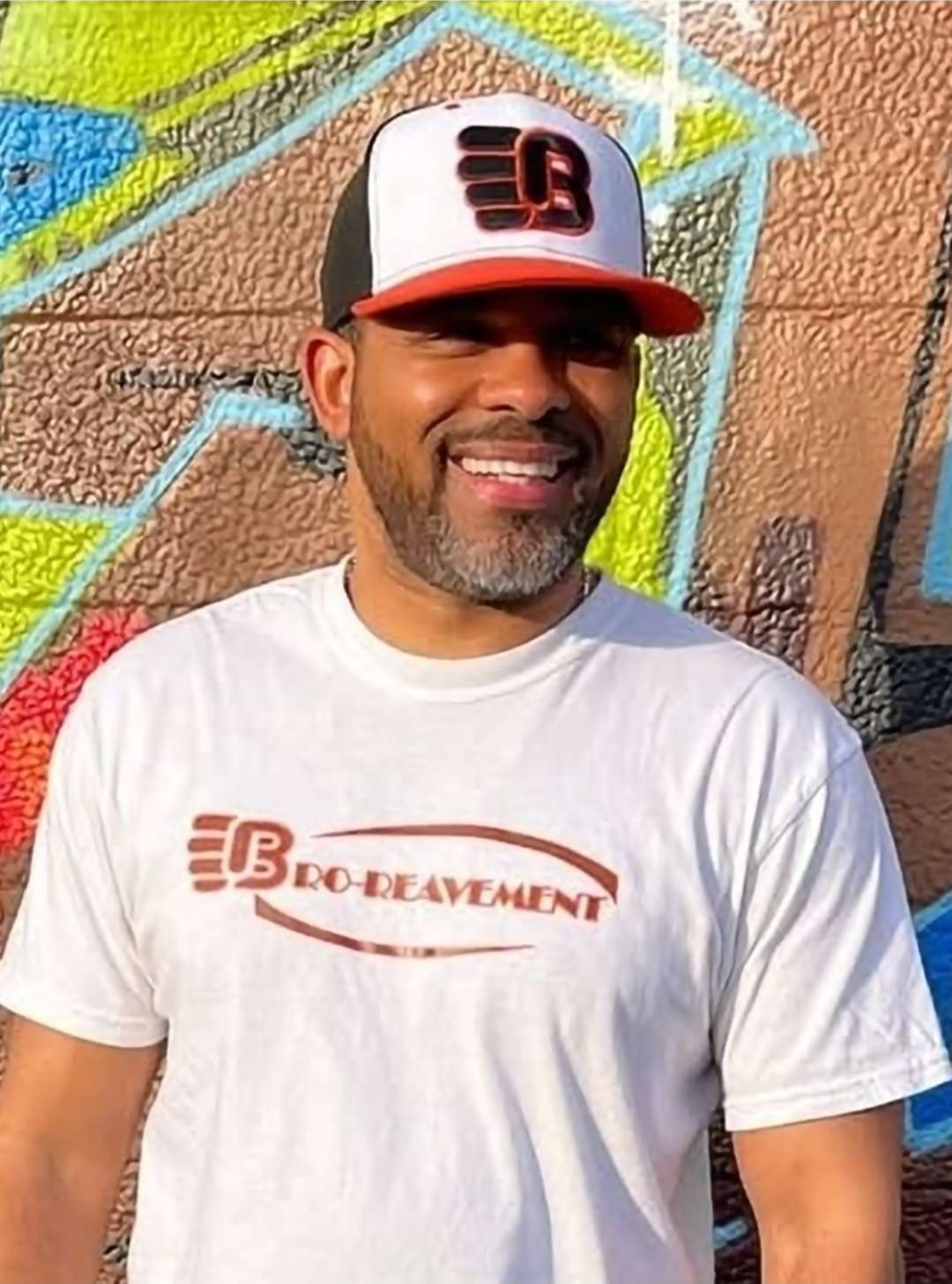 Smiling man wearing a white t-shirt and a black, white, and red cap, standing in front of a colorful graffiti wall.