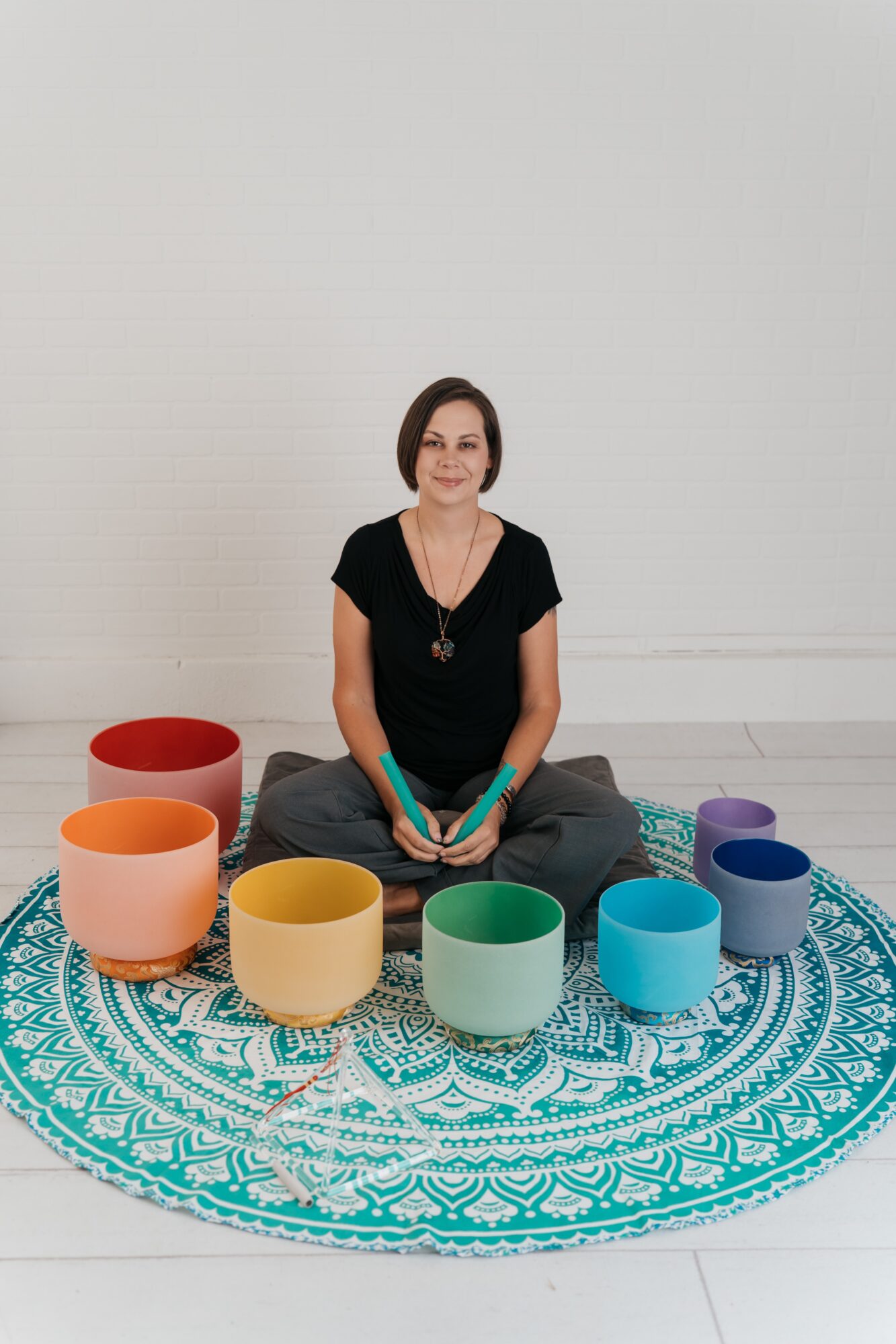 Woman sitting cross-legged on a patterned round rug, surrounded by colorful bowls, holding a book, against a plain white wall.