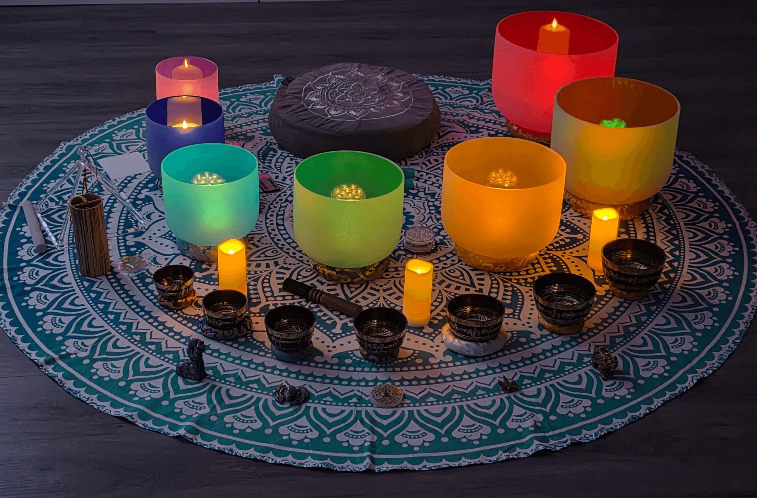 Colorful singing bowls and candles arranged on a patterned round cloth, with a central cushion, on a dark surface.
