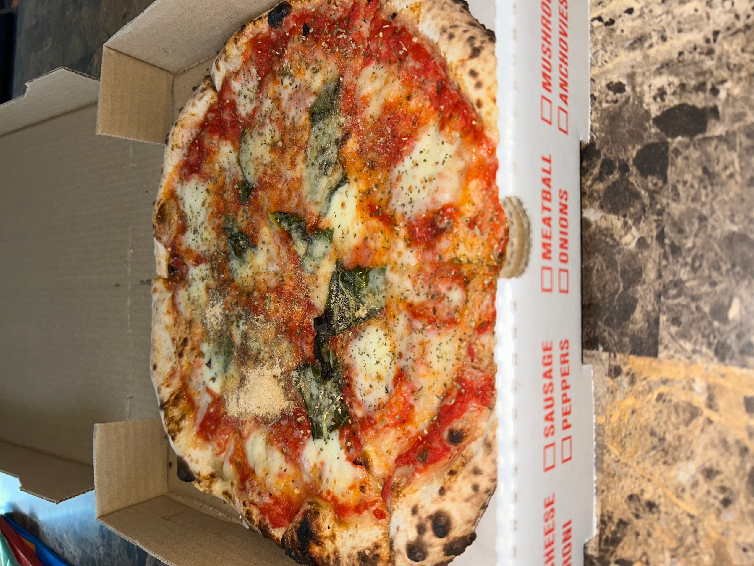 Pizza with cheese, tomato sauce, and basil leaves in an open box on a marble surface.