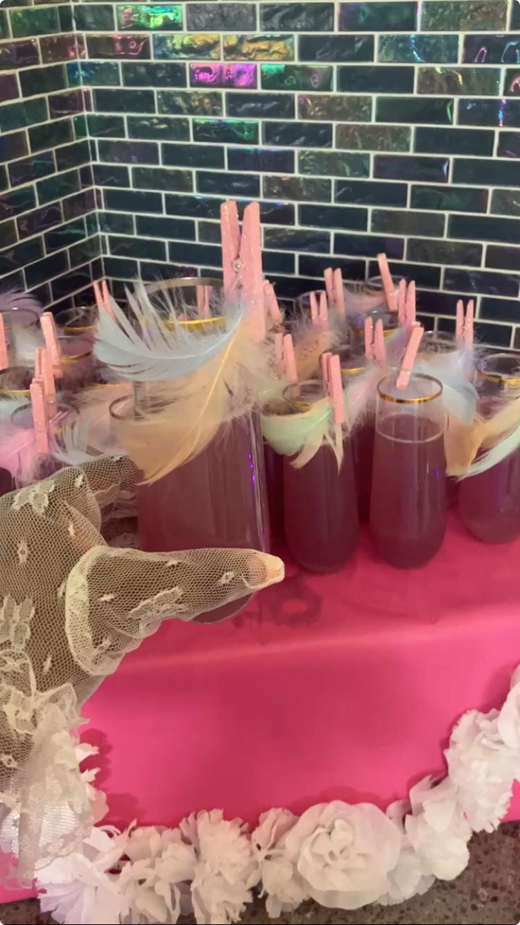 Multiple glasses with pink drinks and garnishes on a pink table, decorated with white flowers, against a brick wall background.
