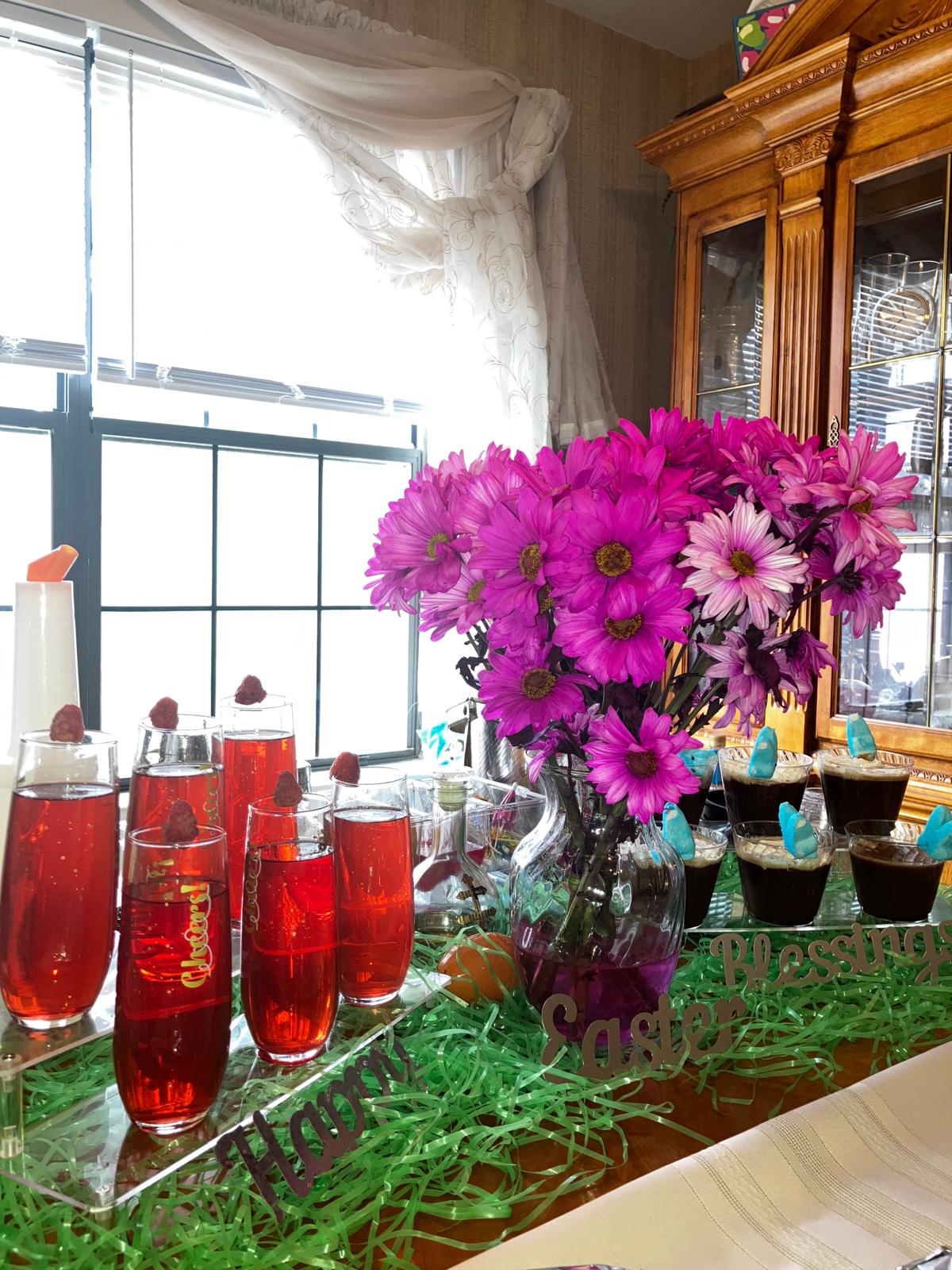 Vase with pink and purple flowers on a table, with red drinks in glasses and a window in the background.