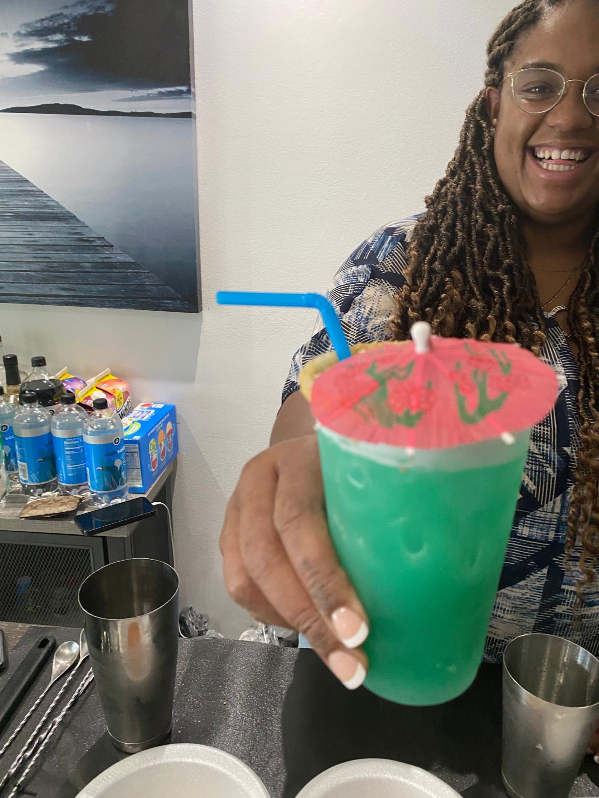 Person holding a green drink with a red umbrella and blue straw, smiling, in a social setting.