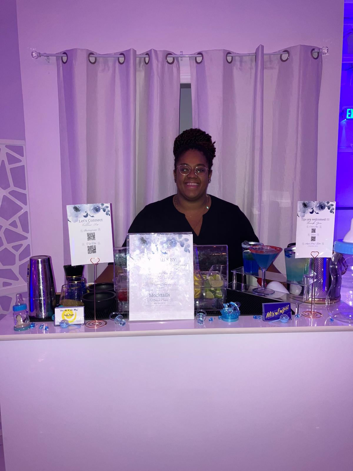 Woman standing behind a bar with drinks and decorations, purple lighting, curtains in the background.