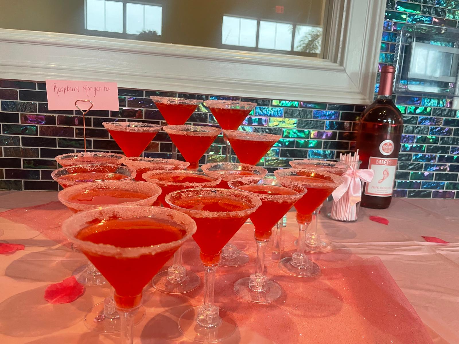 Multiple red cocktails in martini glasses arranged on a table with a pink tablecloth, a bottle, and a window in the background.
