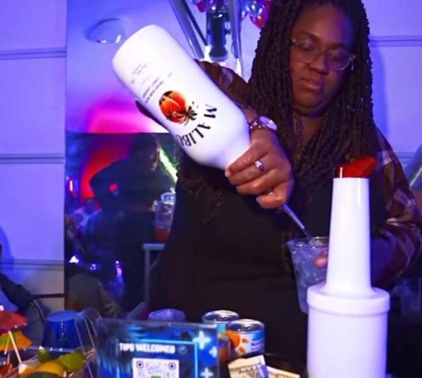 Woman with glasses pours liquid into a glass at a party, colorful lights in background, various items on table.