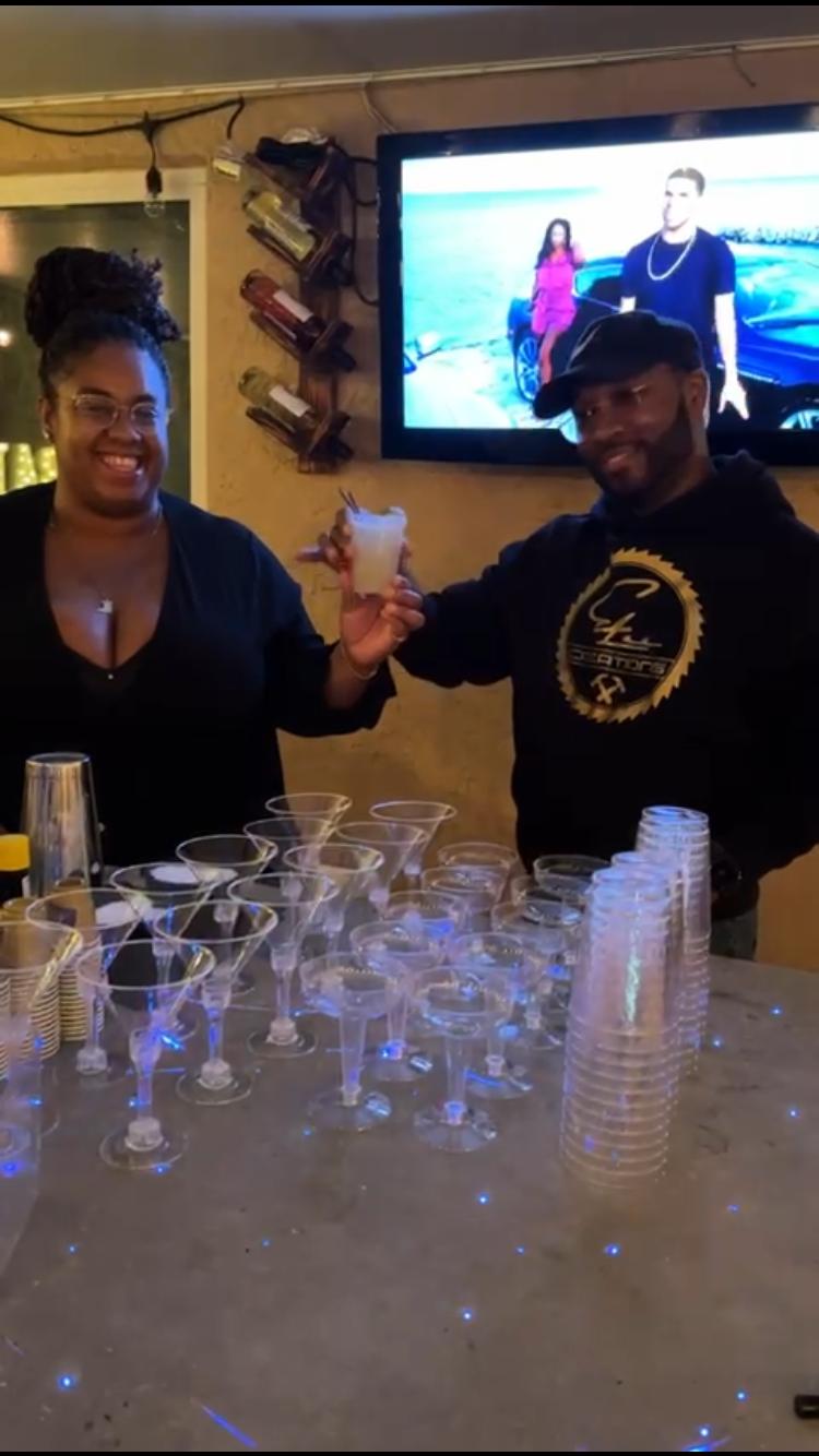 Two people smiling and holding a drink in a bar with a TV screen behind them, glasses on the counter.