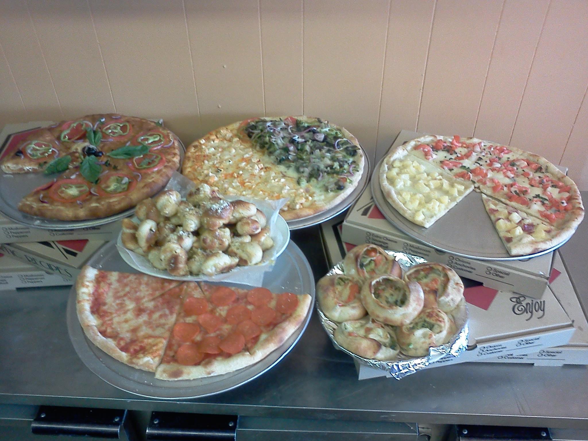 Six pizzas with various toppings on trays, some with sliced pieces missing, on a table against a wall.