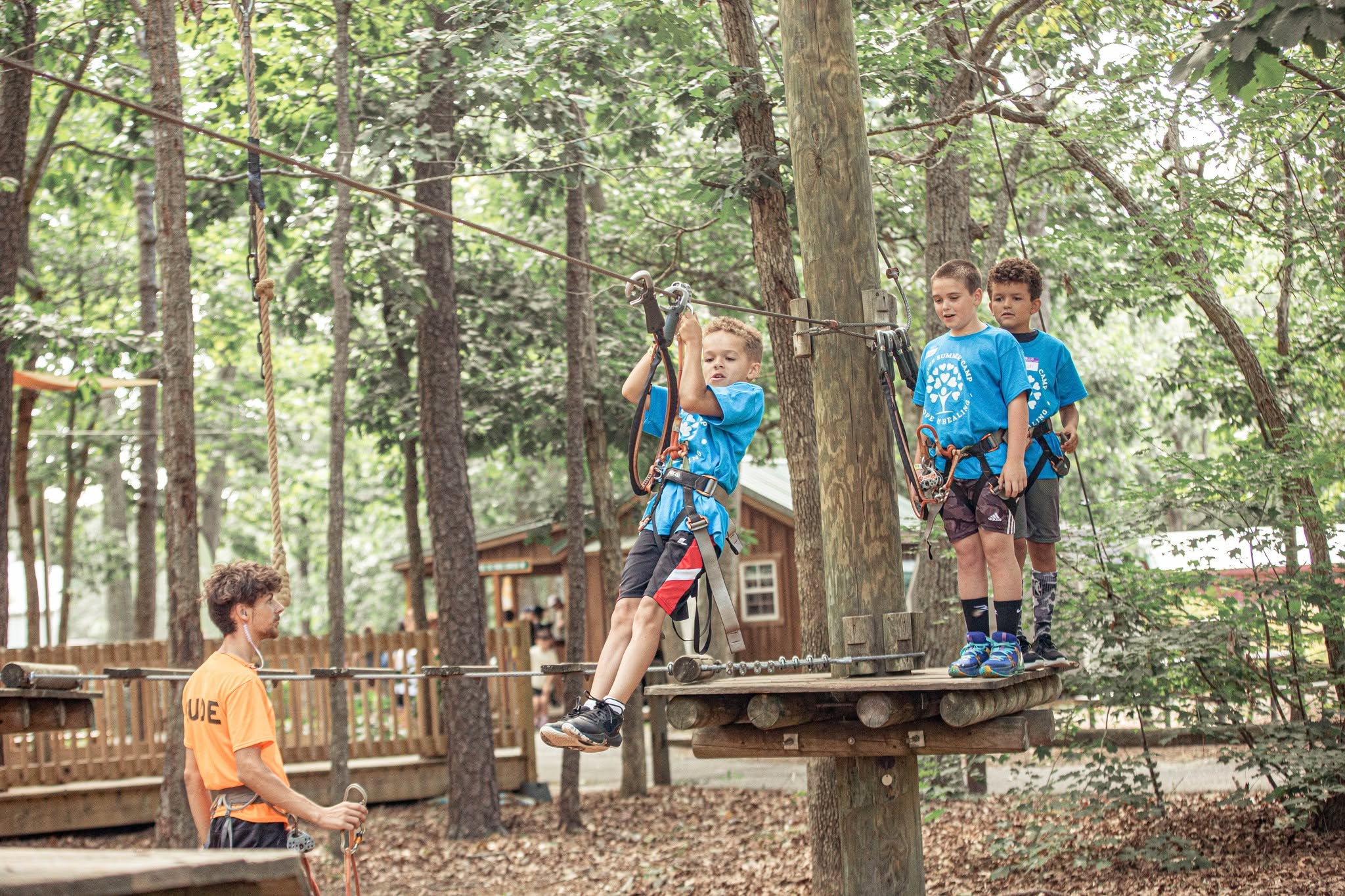 Children participating in a ropes course in a wooded outdoor area, with one child climbing a rope and others waiting on a platform.