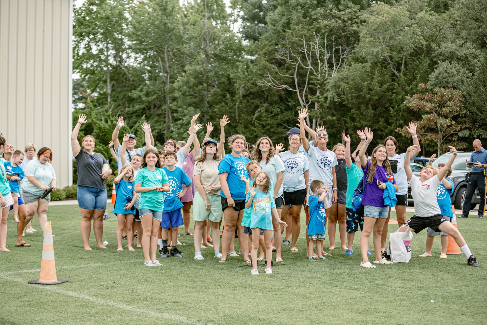 Group of children and adults outdoors, some raising hands, on grassy area with trees in background.