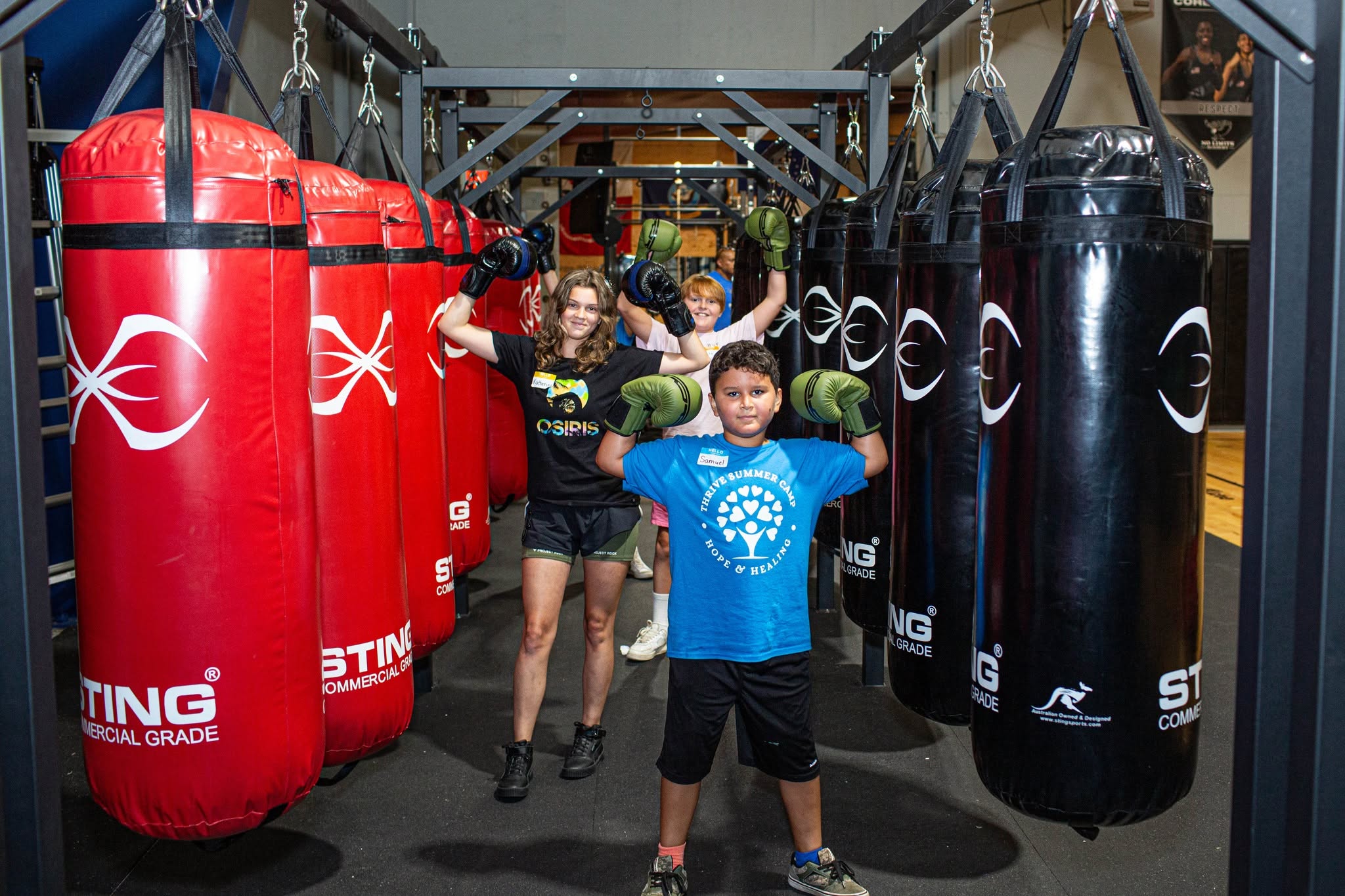 Two children and an adult in a gym with punching bags, children flexing muscles, smiling, and wearing casual clothes.