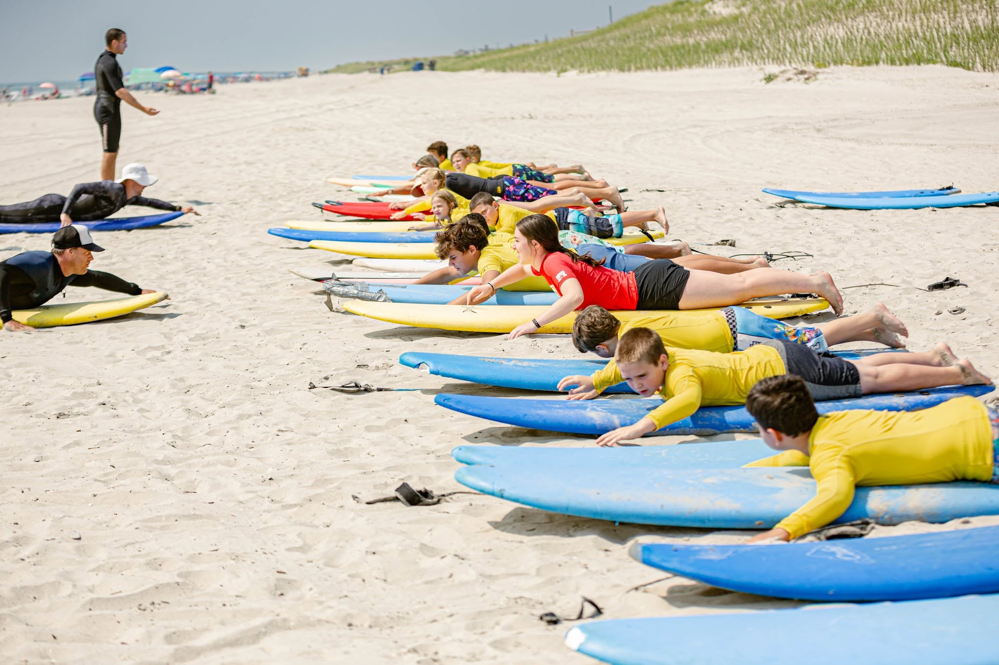 Children in yellow shirts practicing yoga on surfboards on sandy beach, instructor standing nearby, clear sky, grassy dunes in background.
