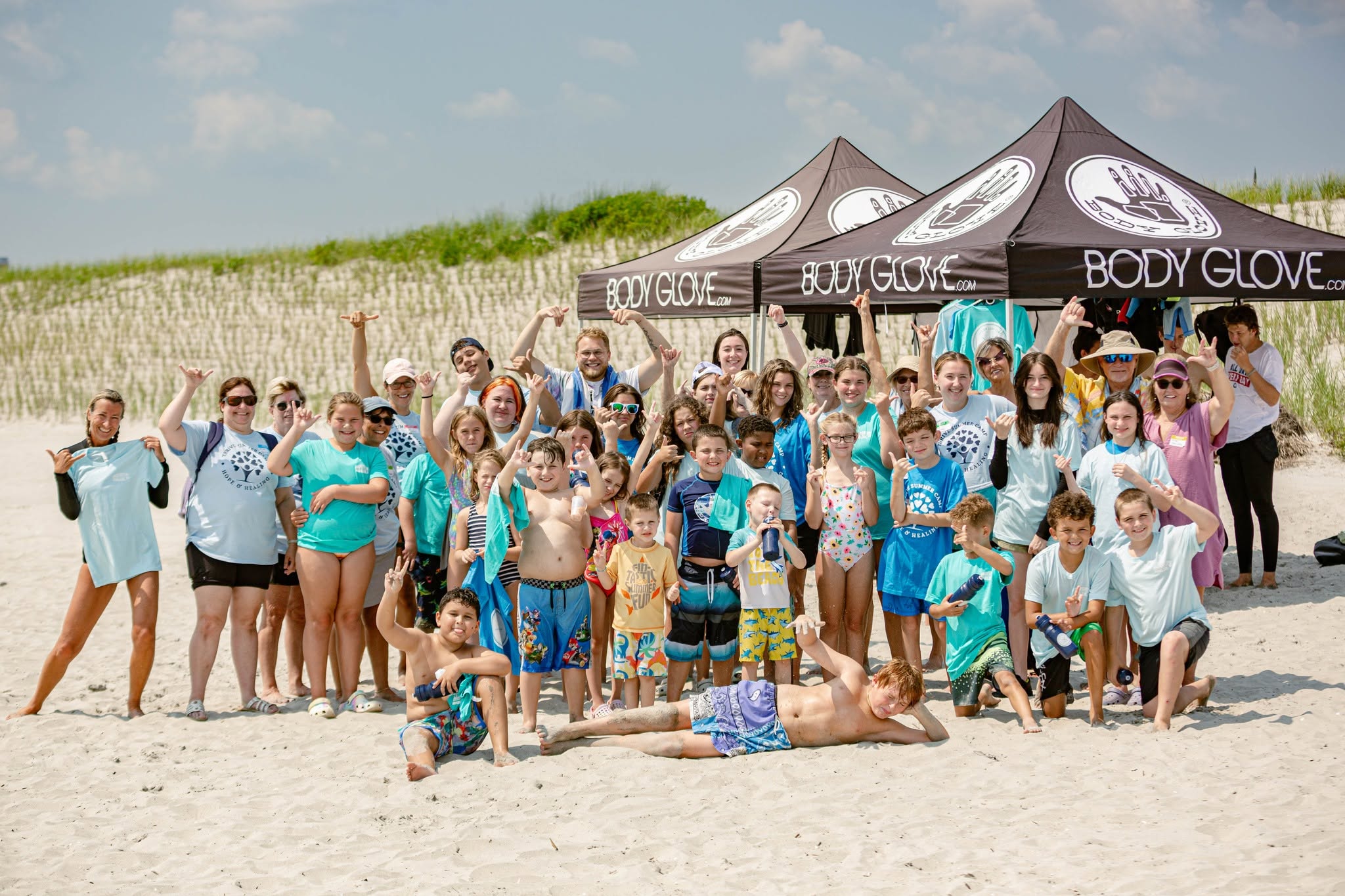 Group of people on a sandy beach in front of tents, some waving, others sitting or lying down, under a blue sky.