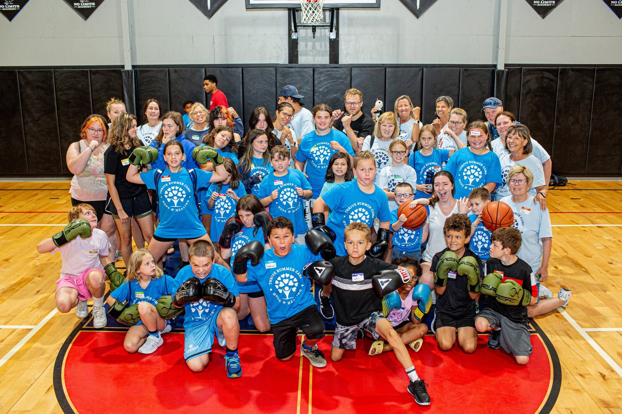Group of children and adults in a gymnasium, some holding basketballs, posing for a photo, with a basketball hoop in the background.