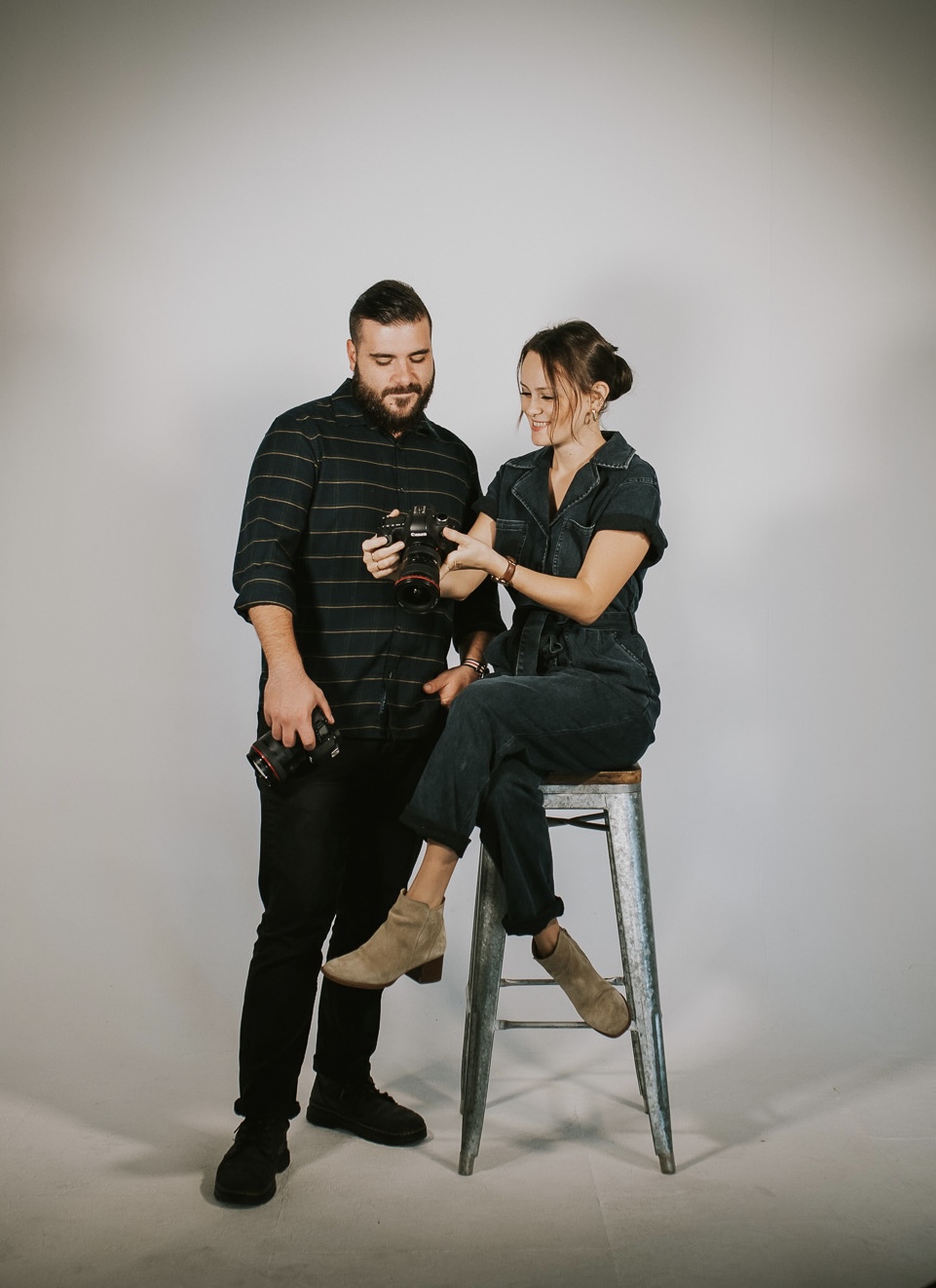 A man and woman stand together, woman sitting on a tall stool, both looking at a phone, against a plain background.