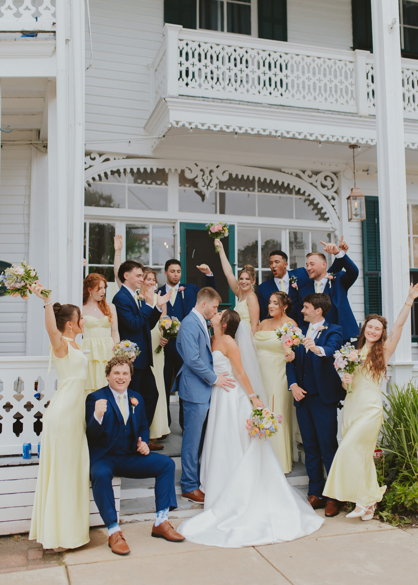 Group of people celebrating wedding outside a white building, some raising hands, holding flowers, and smiling.