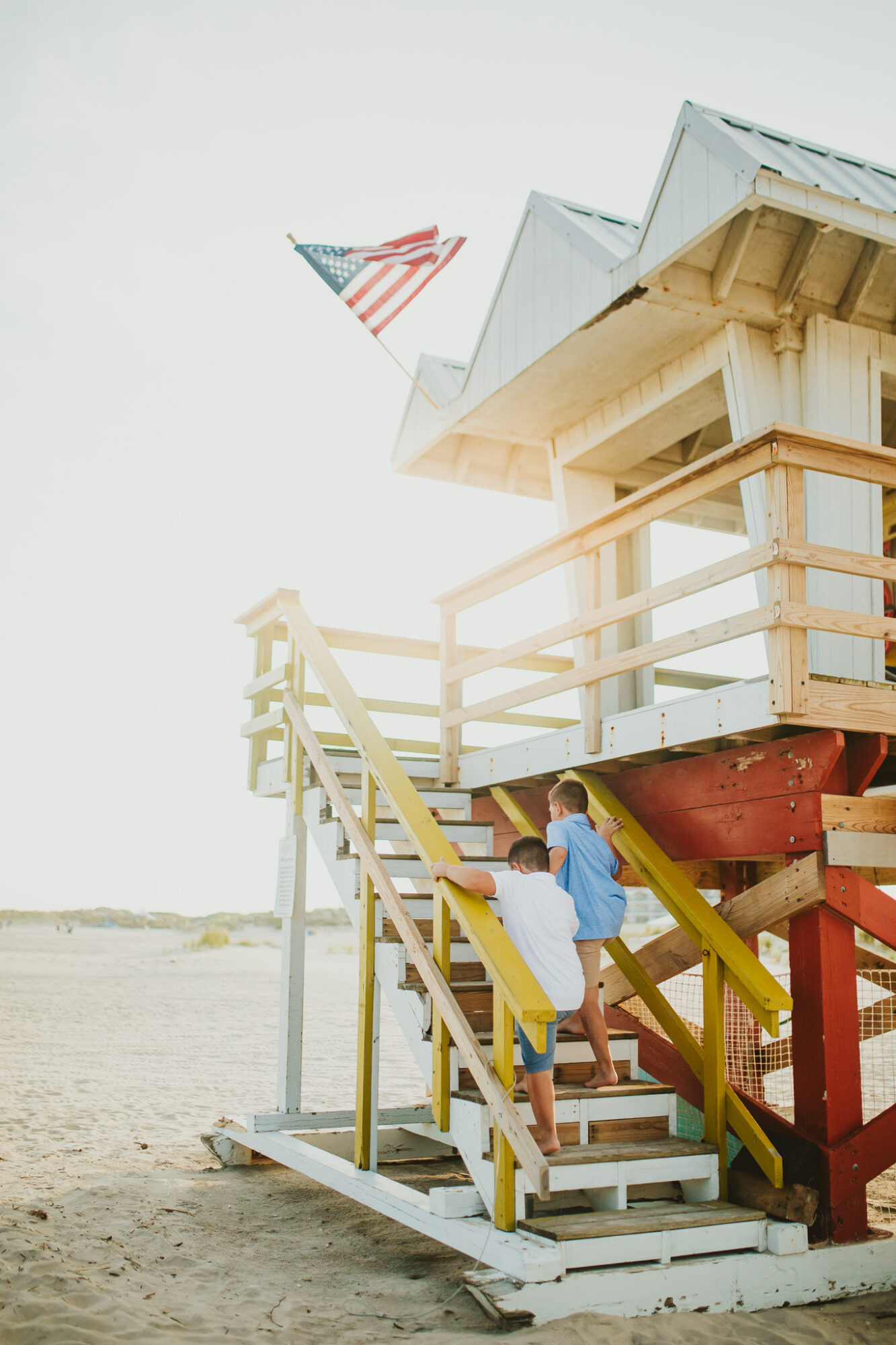 Person with a backpack walking up stairs to a beach lifeguard tower with an American flag, sandy beach, and sky in background.