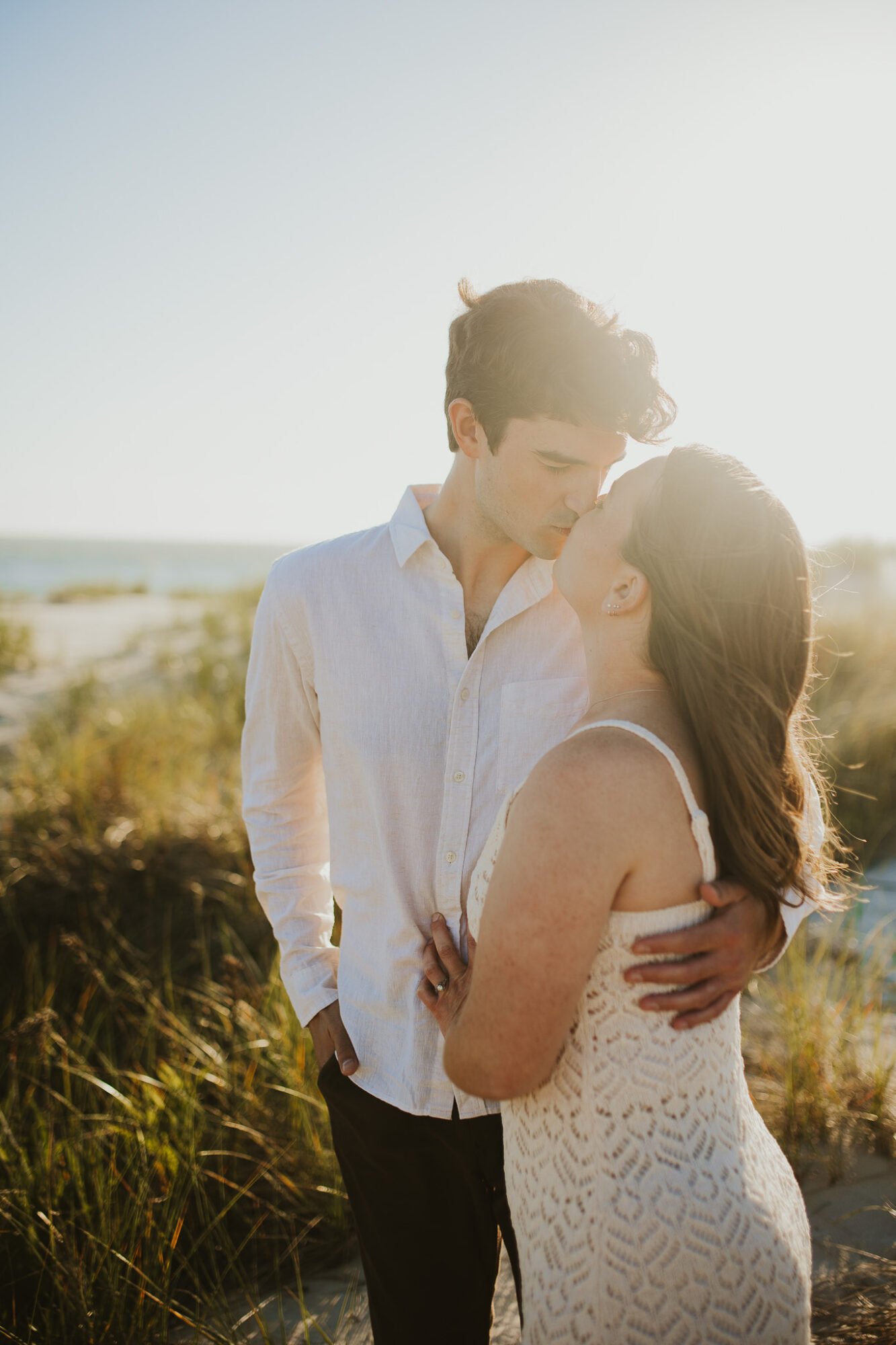 A couple stands outdoors in sunlight, embracing each other with a natural landscape in the background.
