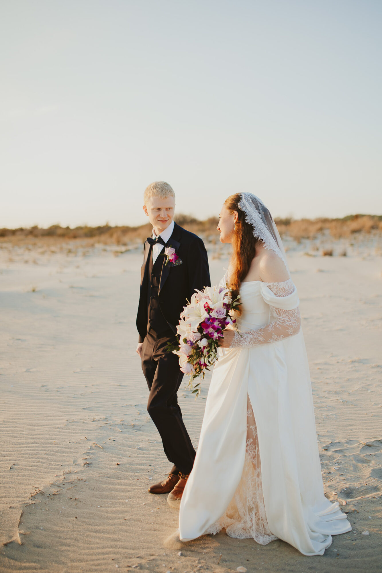 Bride and groom standing on a sandy beach, facing each other, with the bride holding a large bouquet of flowers, under a clear sky.