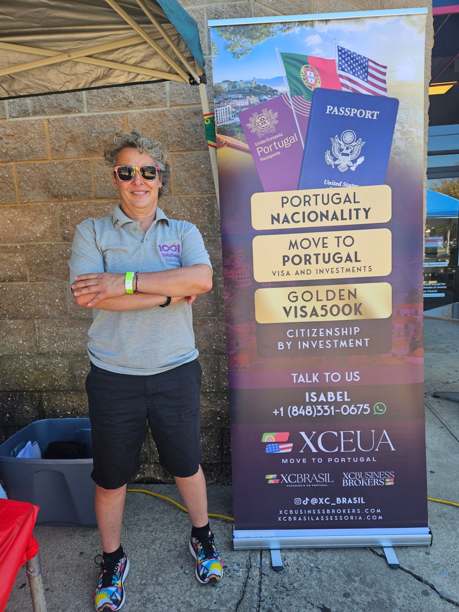 Person standing outdoors next to a tall travel information sign, wearing sunglasses, gray shirt, black shorts, and colorful shoes.