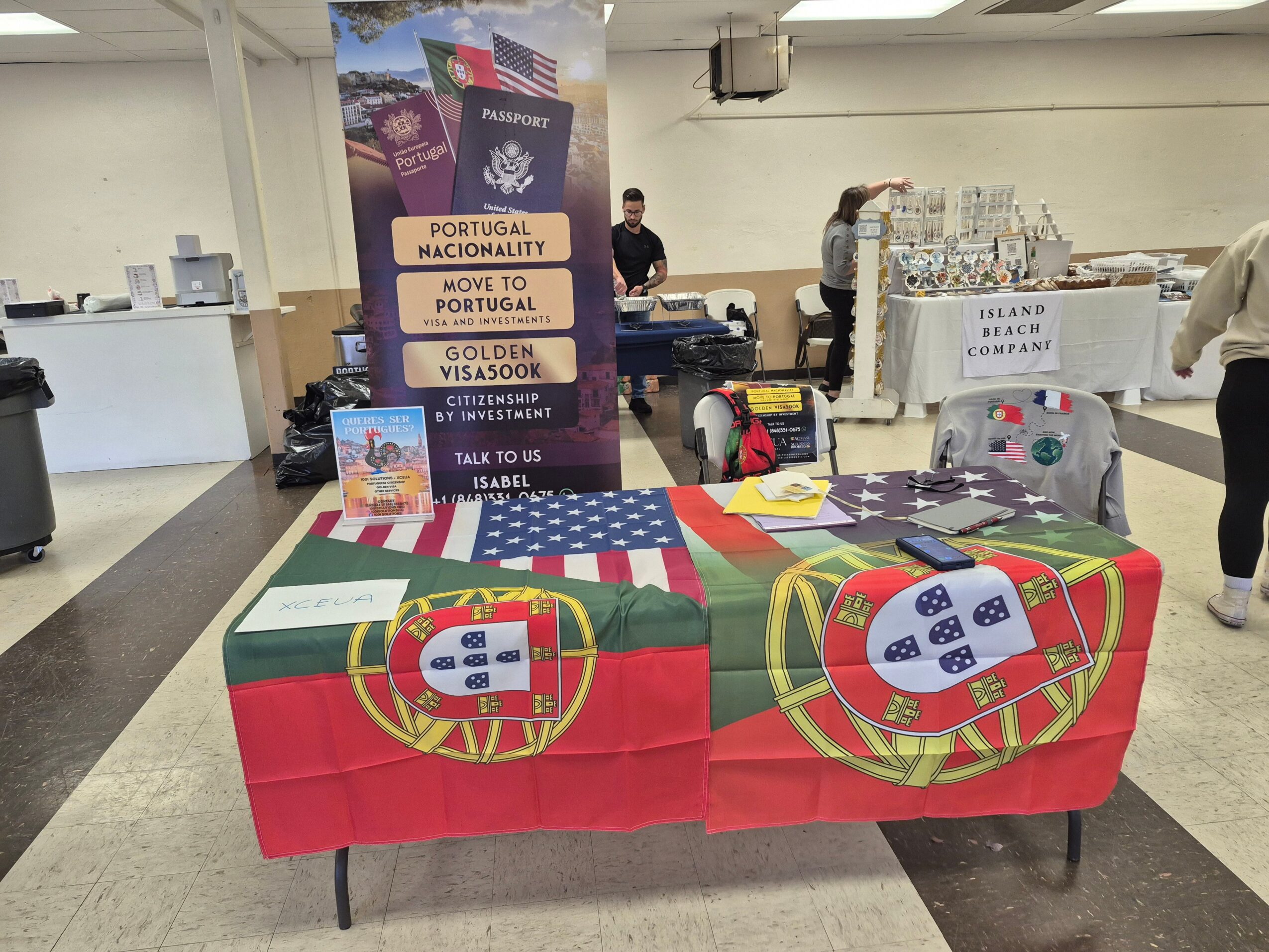 Table covered with Portuguese flag, with informational materials and a banner behind it, in an indoor setting.