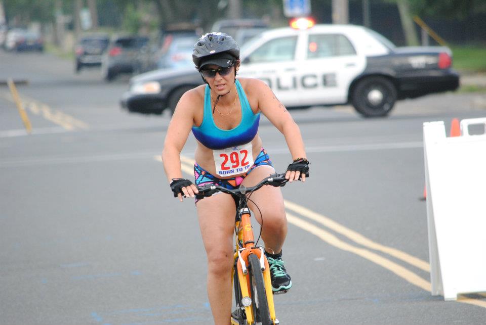 Female cyclist wearing sunglasses, helmet, and blue top riding a bike on a street with police car in background
