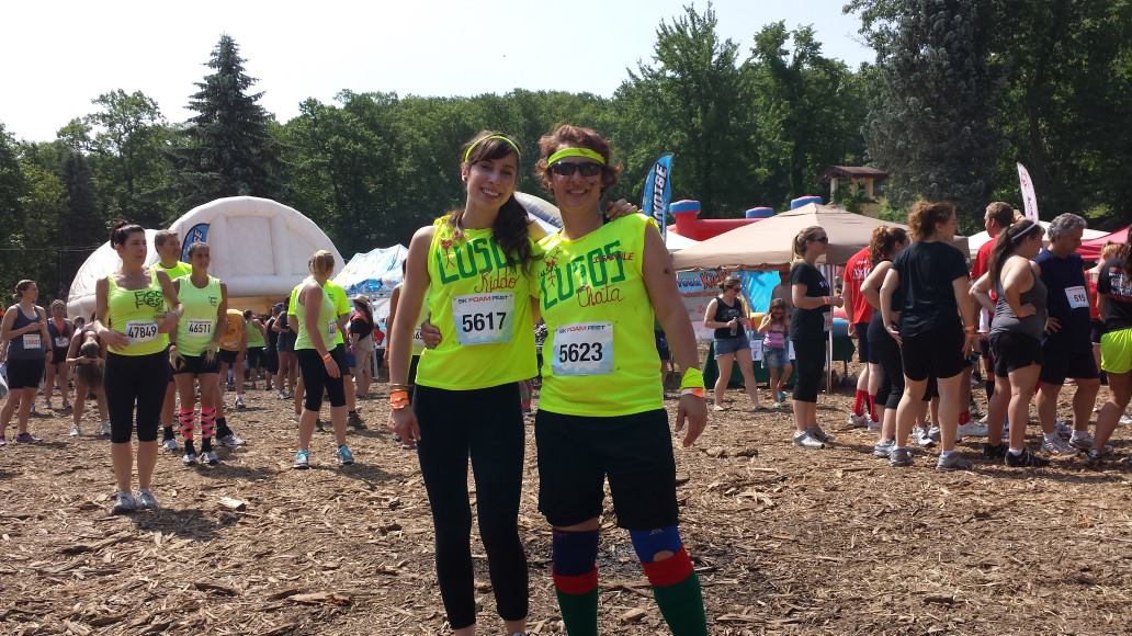 Two people in bright yellow shirts and race bibs stand outdoors among a crowd, trees, and tents in the background.