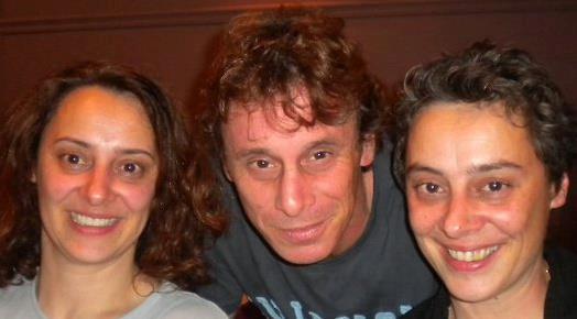 Three people smiling closely together, two women and one man, in an indoor setting.