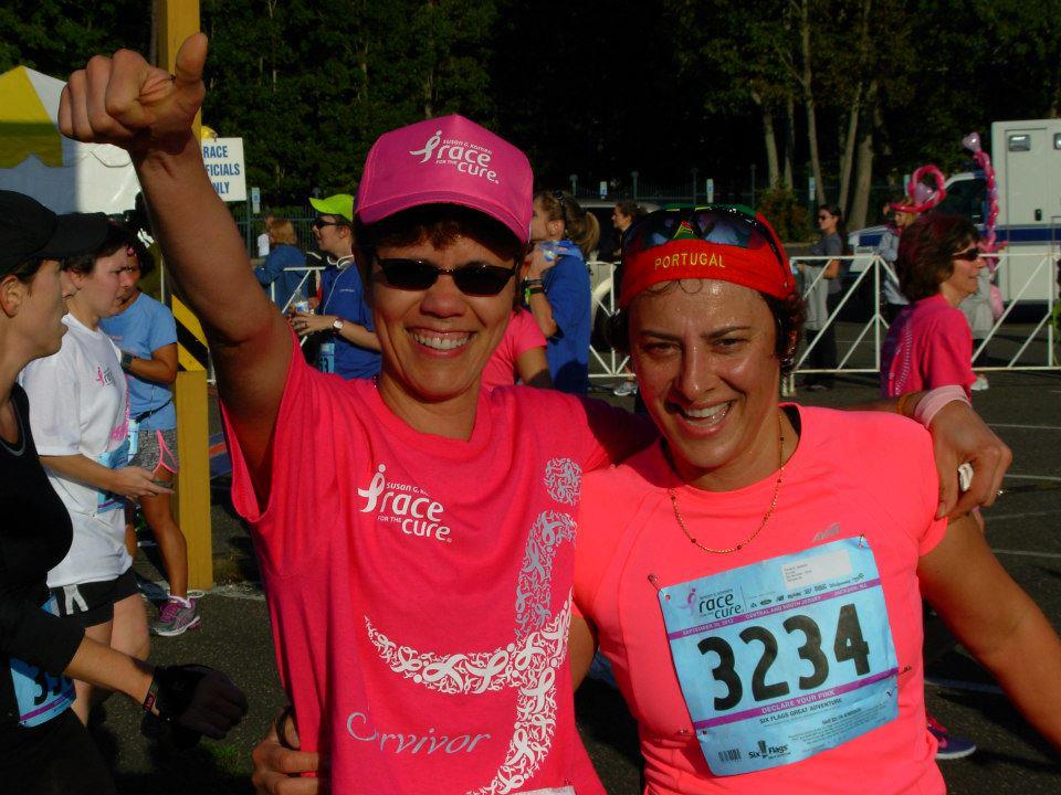 Two women in pink shirts and hats smiling, celebrating at a race event with other participants in the background.
