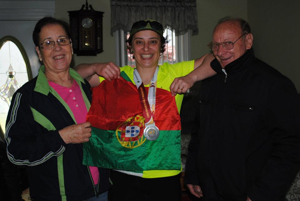 Three people smiling, one holding a flag and medals, in a room with a clock and window behind them.