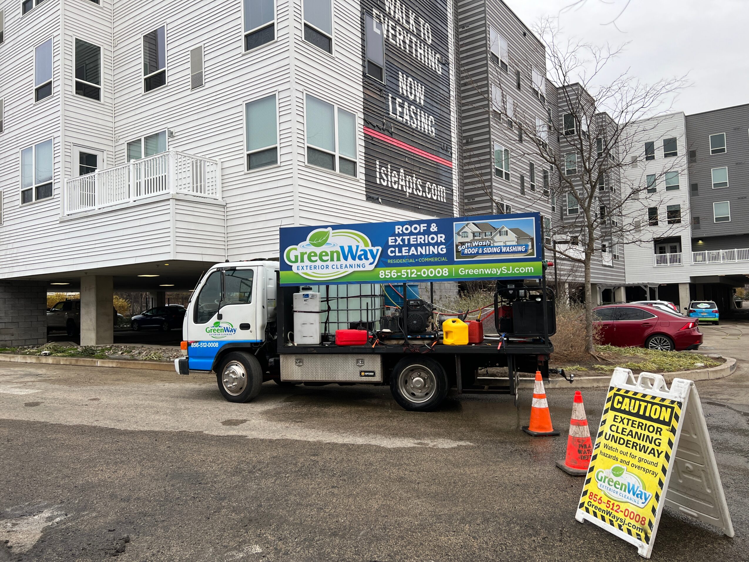 Service truck parked on street with caution sign and traffic cones, buildings in background.
