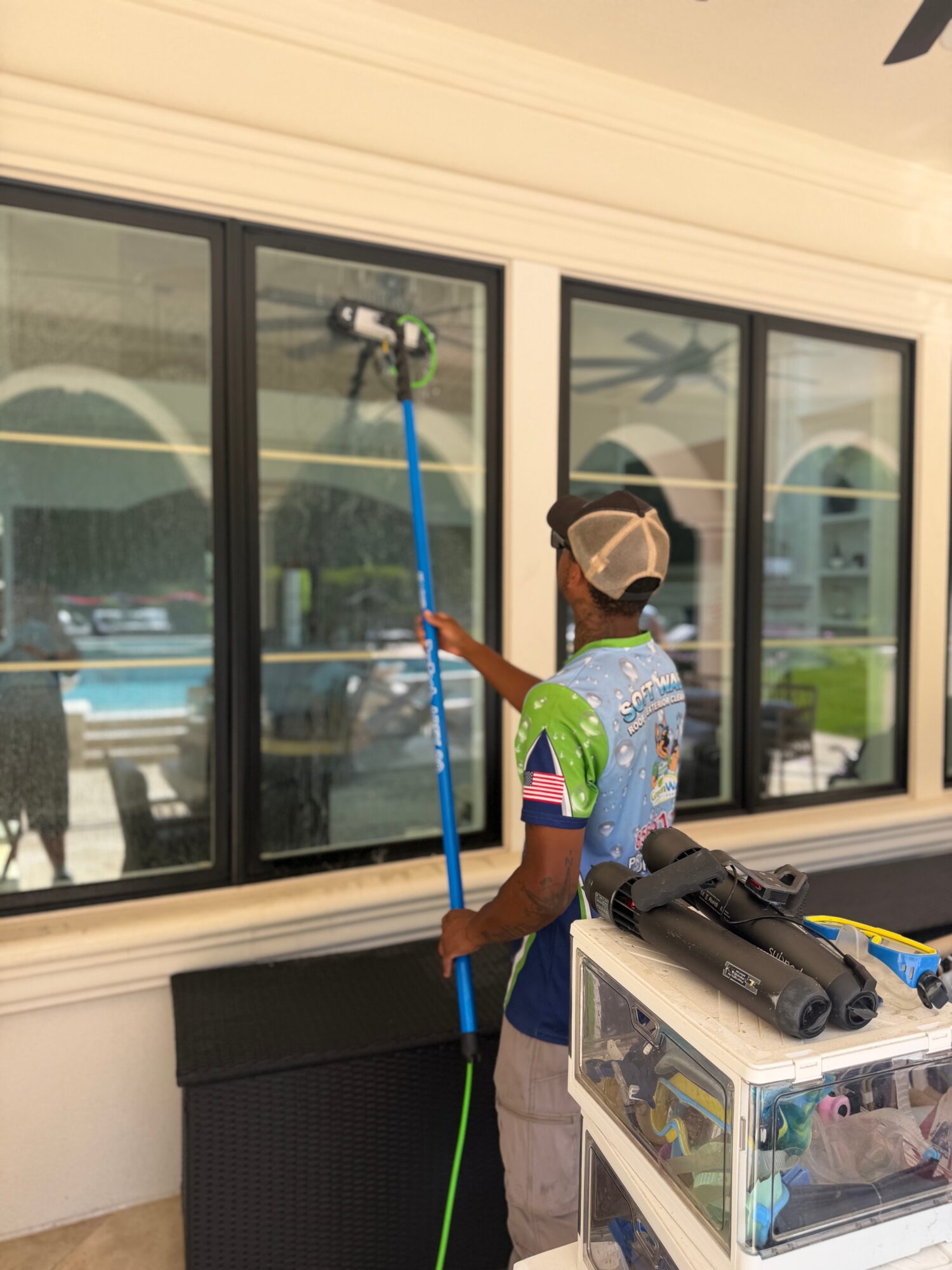 Child cleaning window with a long-handled tool, wearing a cap and colorful shirt, near a white wall and black furniture.