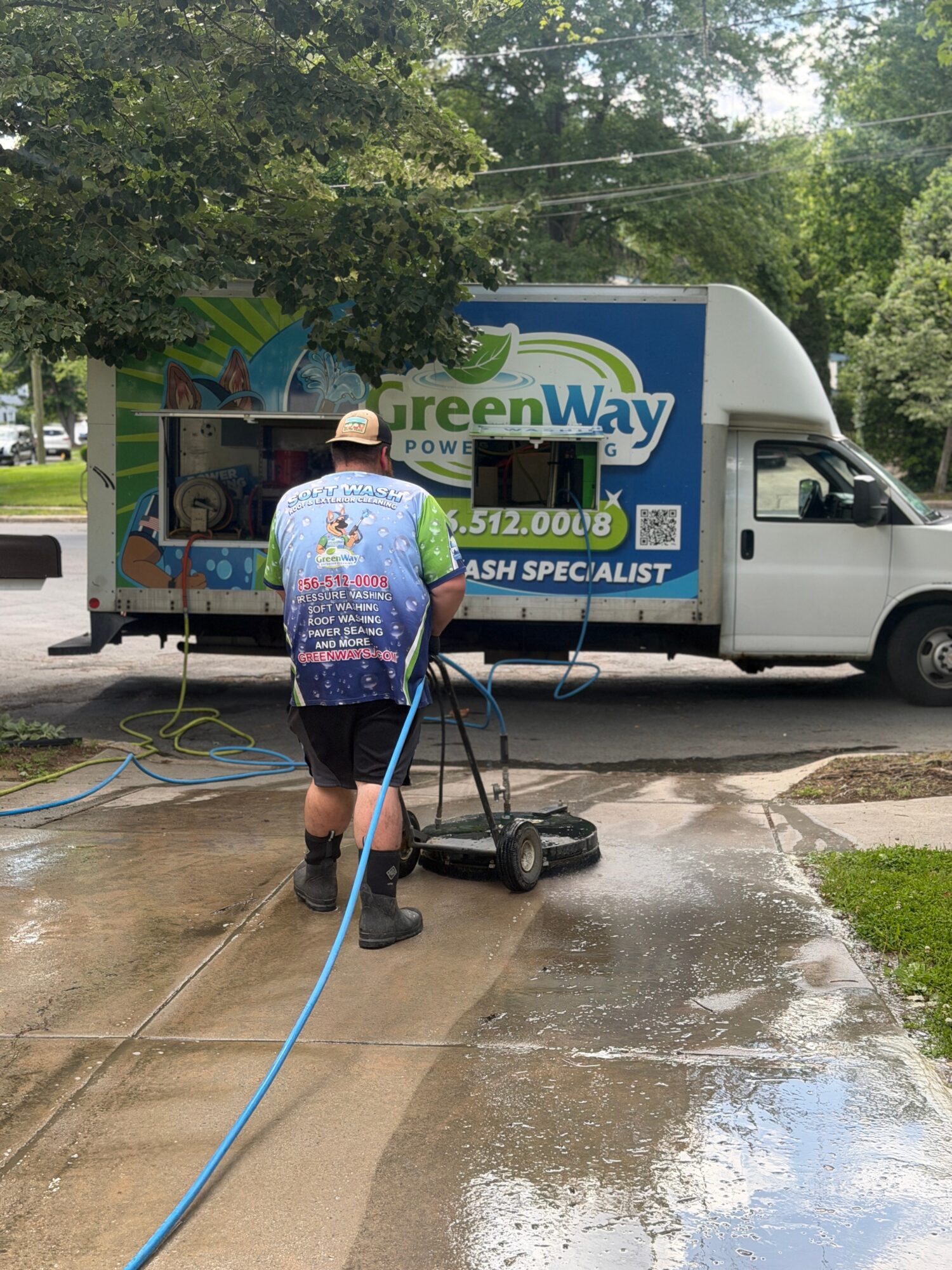 Person cleaning a vehicle wash station with a GreenWay mobile truck in a park-like area, trees and grass visible.