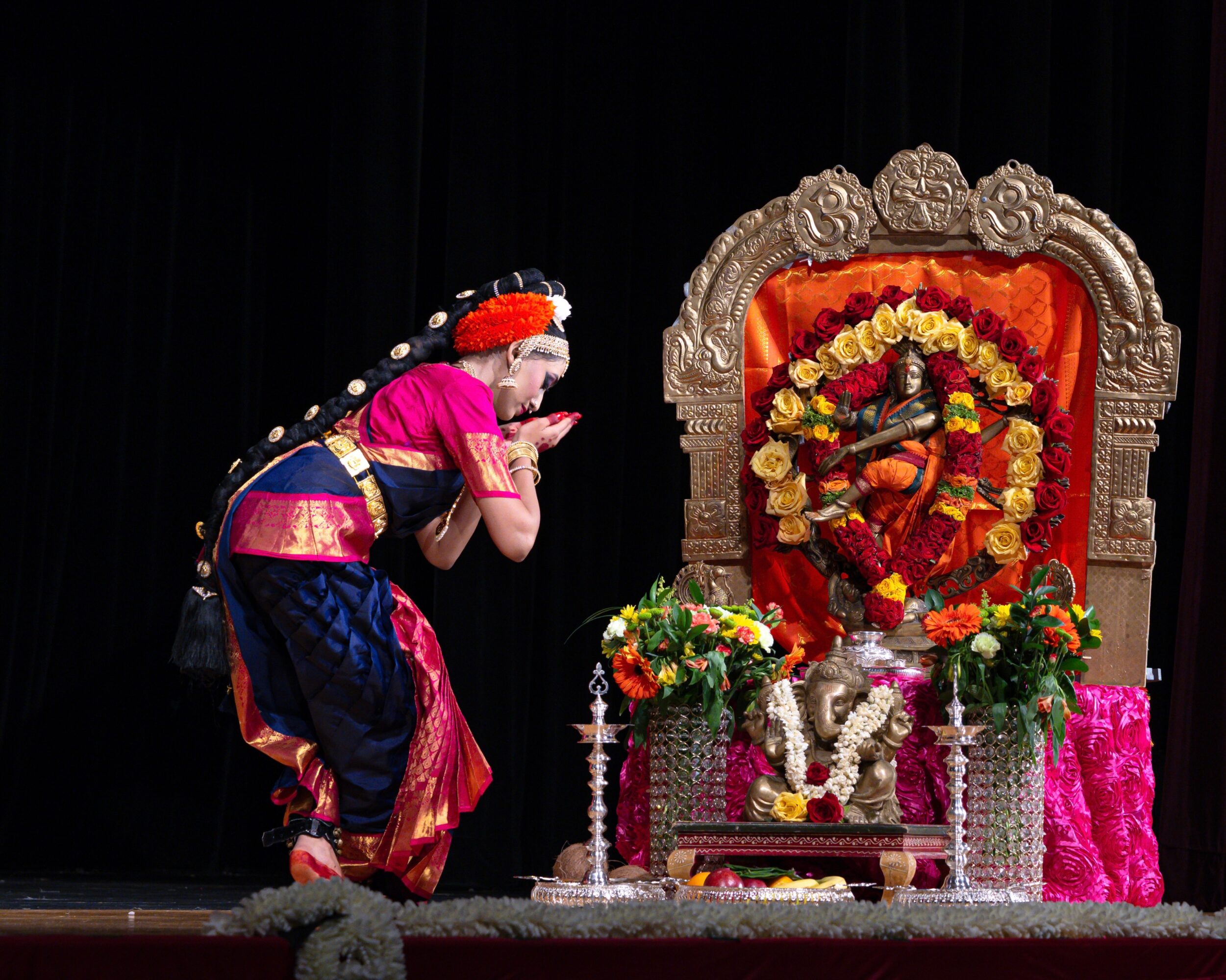 Person in traditional attire performing a prayer before a decorated idol on stage.