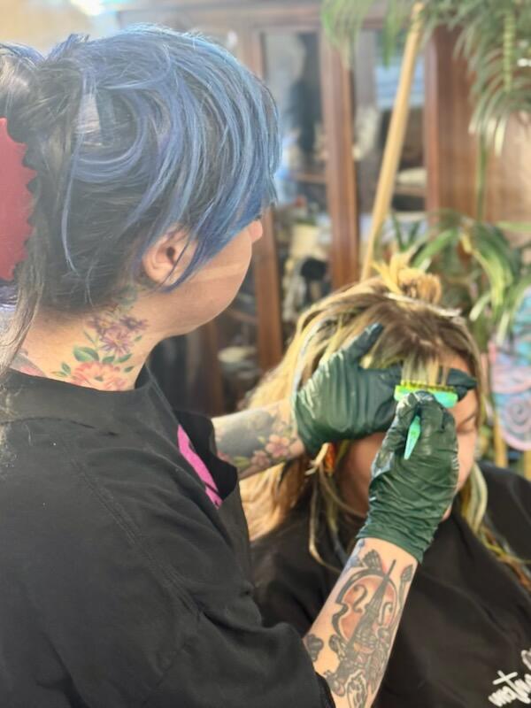 Tattooed person with blue hair tattooing a person's head in a salon, surrounded by plants and salon equipment.