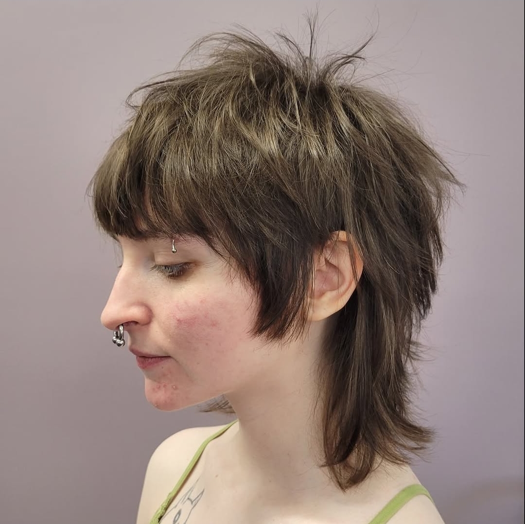 Side view of a person with a layered, messy hairstyle and a septum piercing, wearing a light green top.