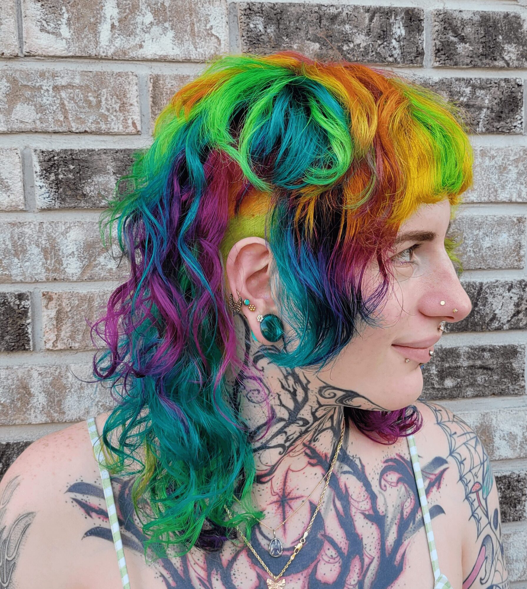 Person with rainbow-colored hair, tattoos on neck and shoulders, and a nose piercing, standing against a brick wall.