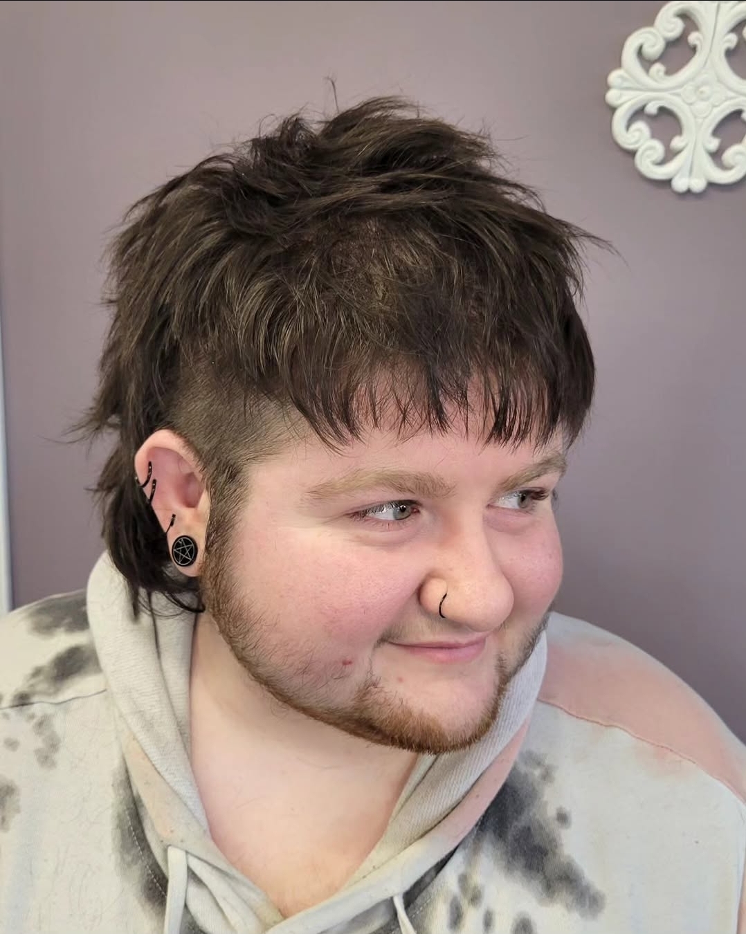 Young person with short, layered hair, multiple earrings, and a nose ring, wearing a hoodie, smiling slightly.