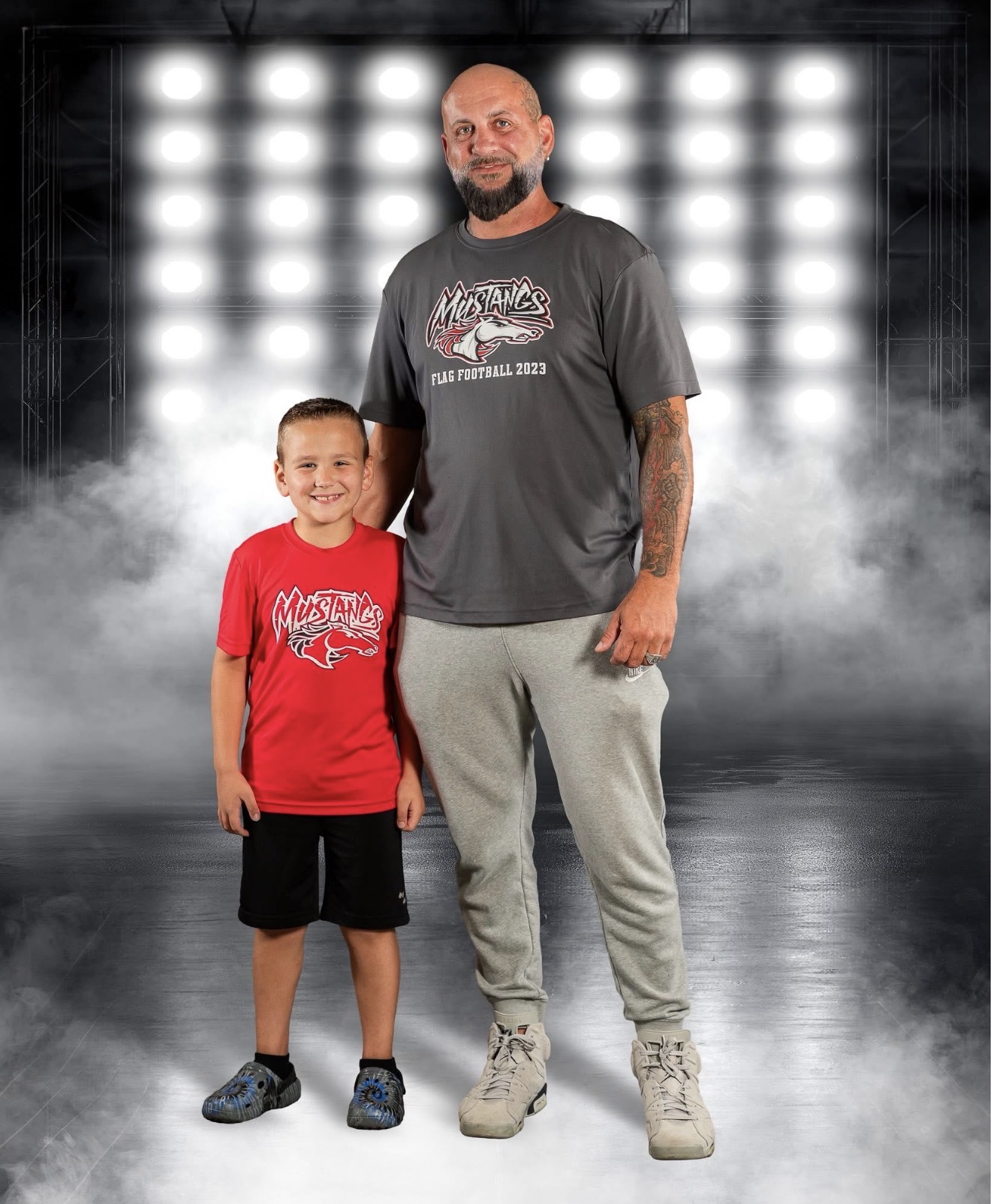 A man and a boy stand together on a stage with bright lights behind them, smiling. The man wears a gray t-shirt and gray pants, the boy wears a red t-shirt and black shorts.