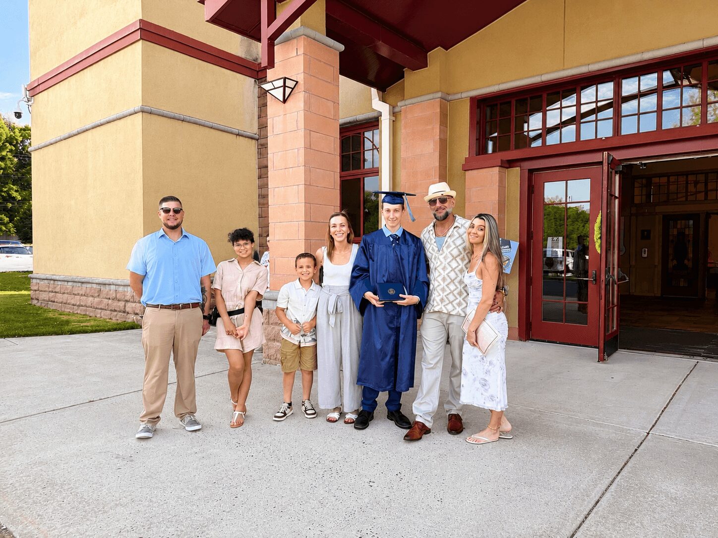 Group of seven people, including a graduate in cap and gown, standing outside a building with yellow and red walls.