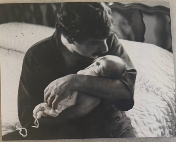Person holding a newborn baby, sitting on a bed, in black and white.
