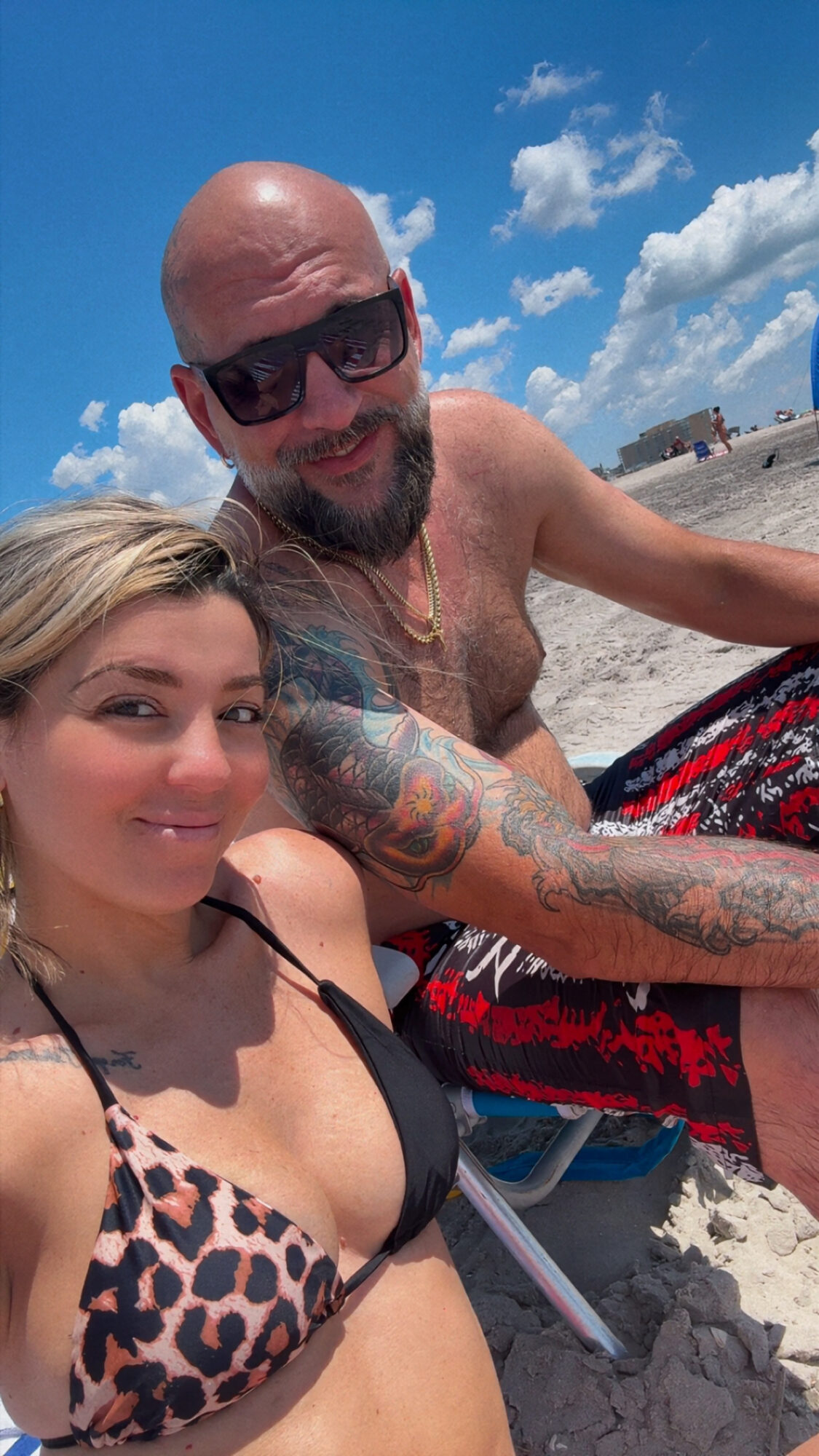 A man and woman sitting on a beach, smiling, with blue sky and clouds in the background.
