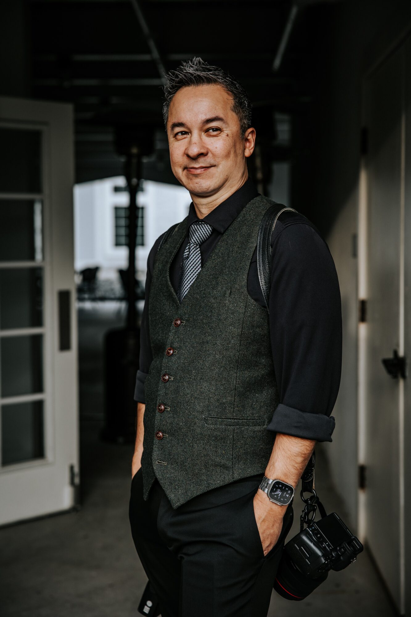 Man with dark hair, wearing a vest, shirt, and tie, holding a camera, standing in an indoor setting.