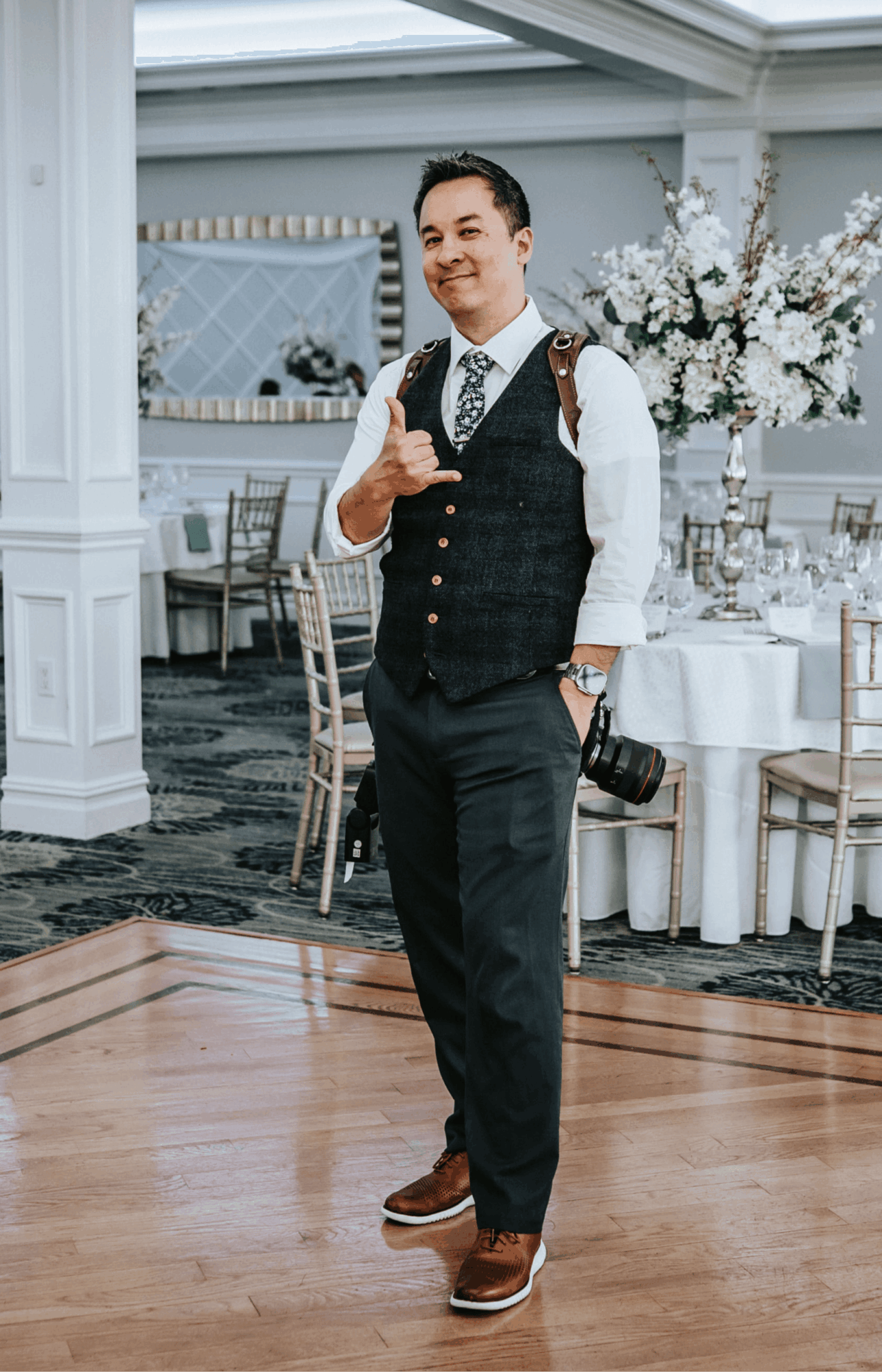 Man in formal attire holding a camera, standing in a decorated event space with tables and floral arrangements.