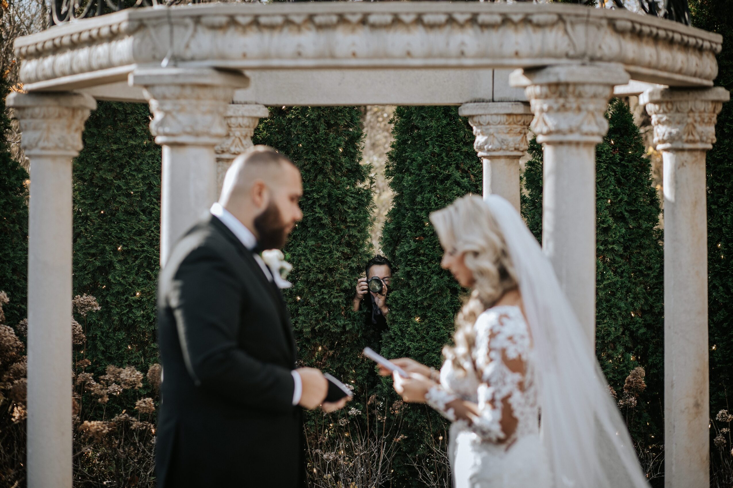 Bride and groom exchanging vows outdoors with a classical gazebo and greenery in the background.